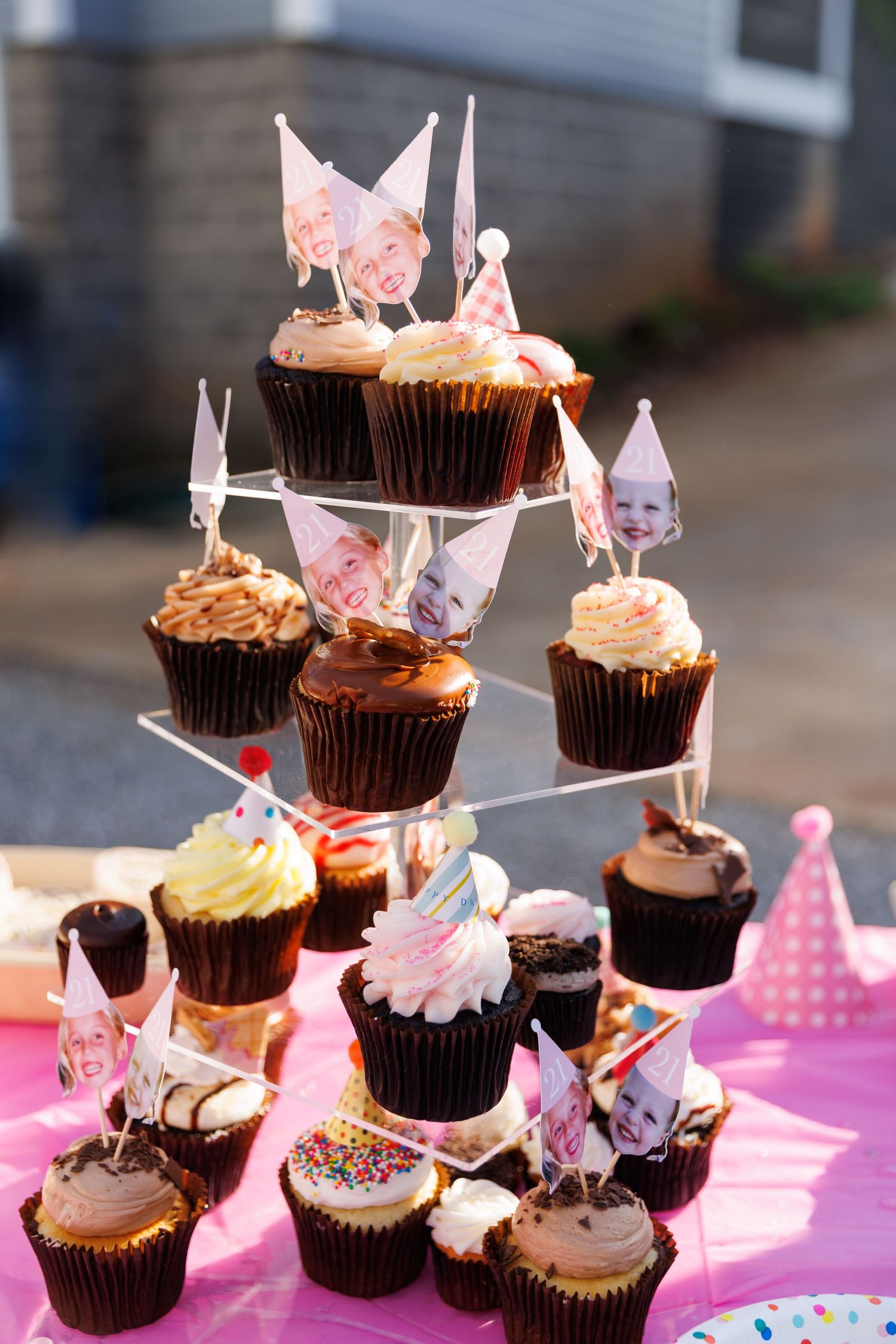 Tiered cupcake stand with chocolate cupcakes, photo toppers, and colorful decorations on a pink table at a birthday party.