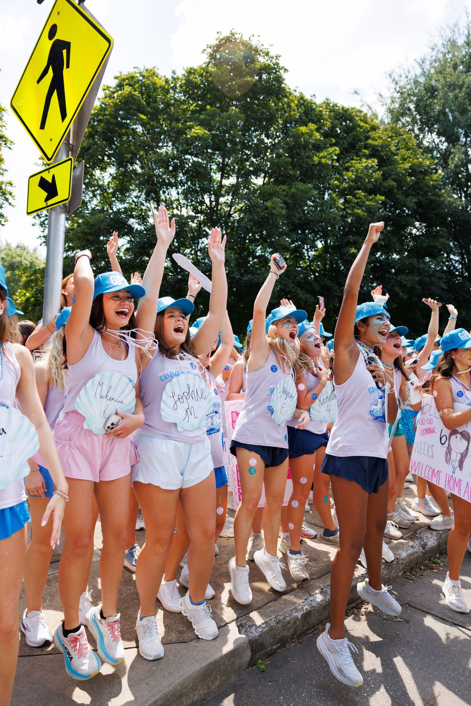 Excited sorority women in blue caps and pink-white outfits cheer with raised arms during Greek recruitment day.
