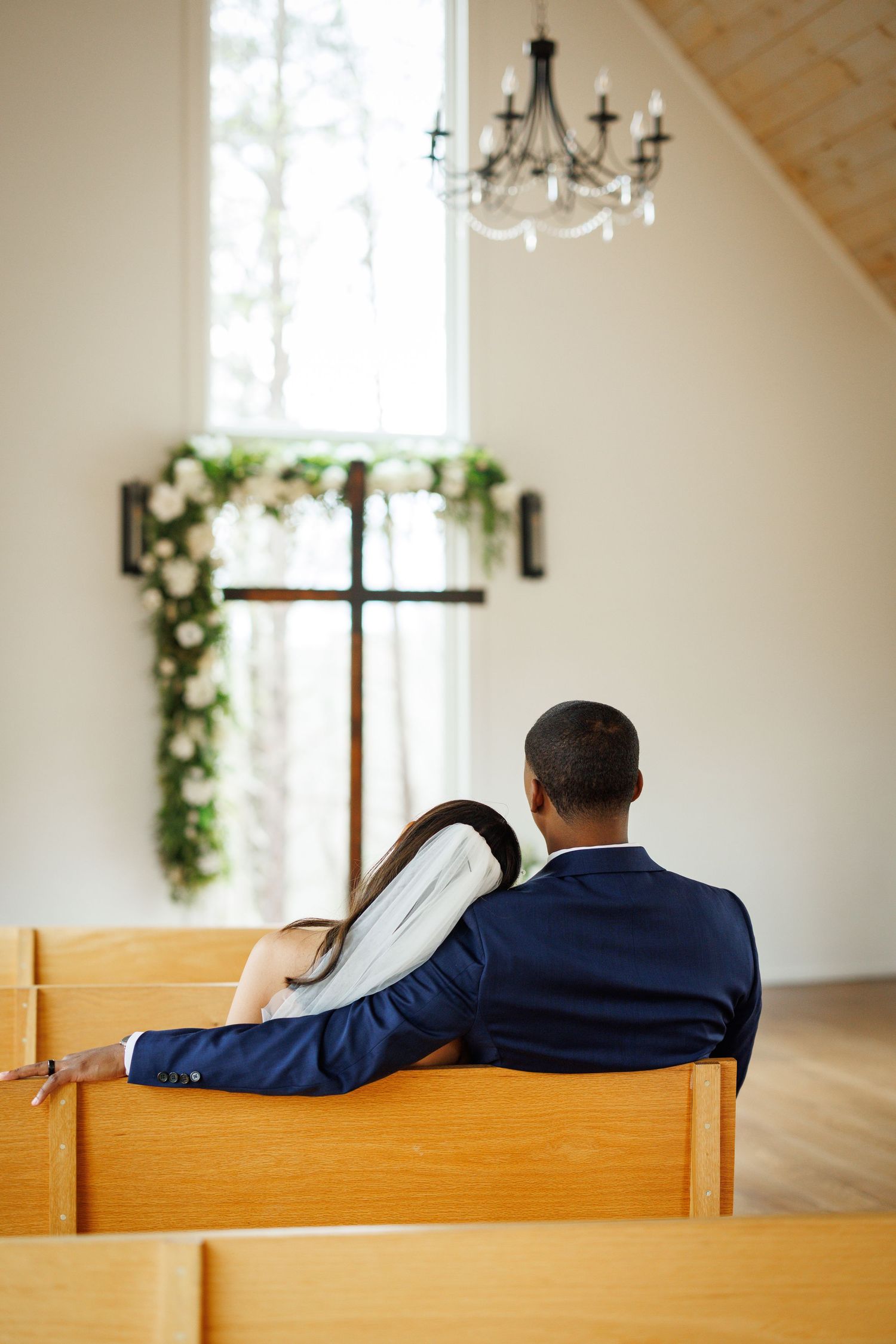 A quiet moment in a chapel with a couple sitting together on a wooden pew beneath a floral-adorned window.