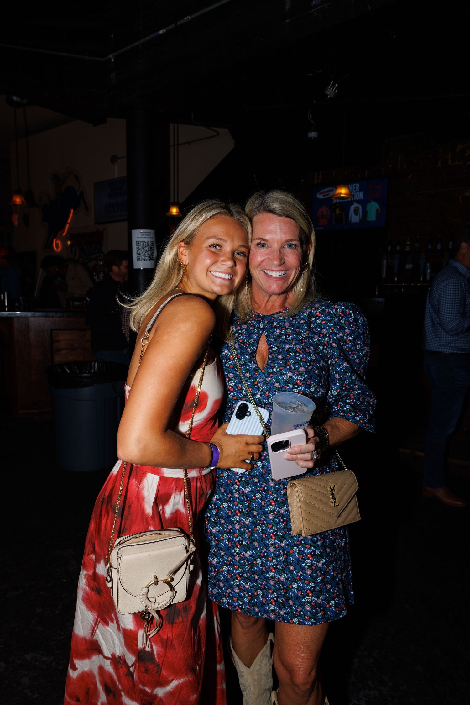 Two smiling women posing together at a nightclub, one in a red floral dress and one in a blue floral mini dress.