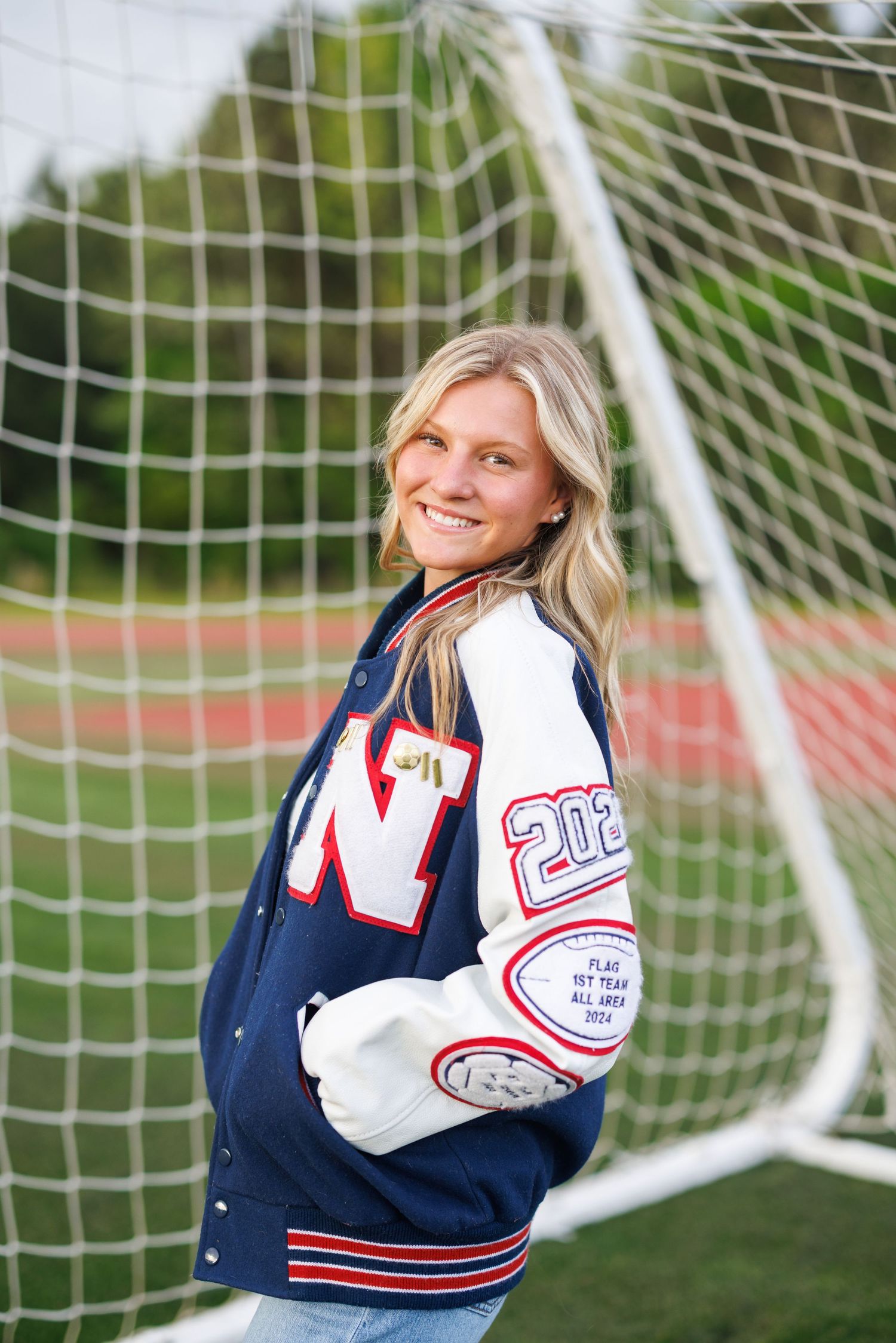 Smiling teen girl in varsity letter jacket poses by soccer goal net on field for senior portrait photo.