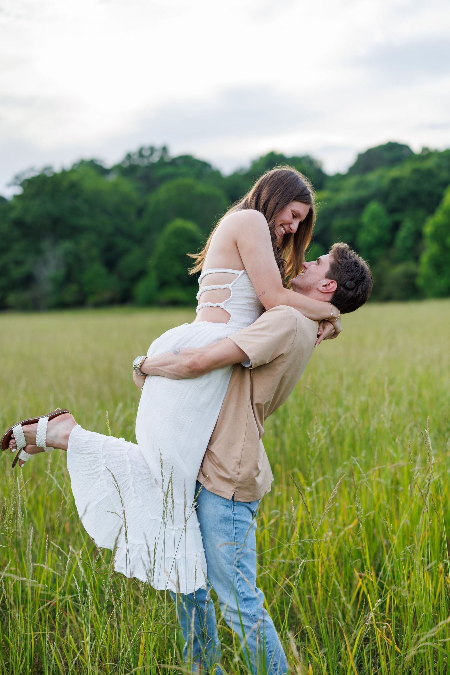 A man lifts a woman in a white dress in a green field, sharing a romantic moment outdoors.