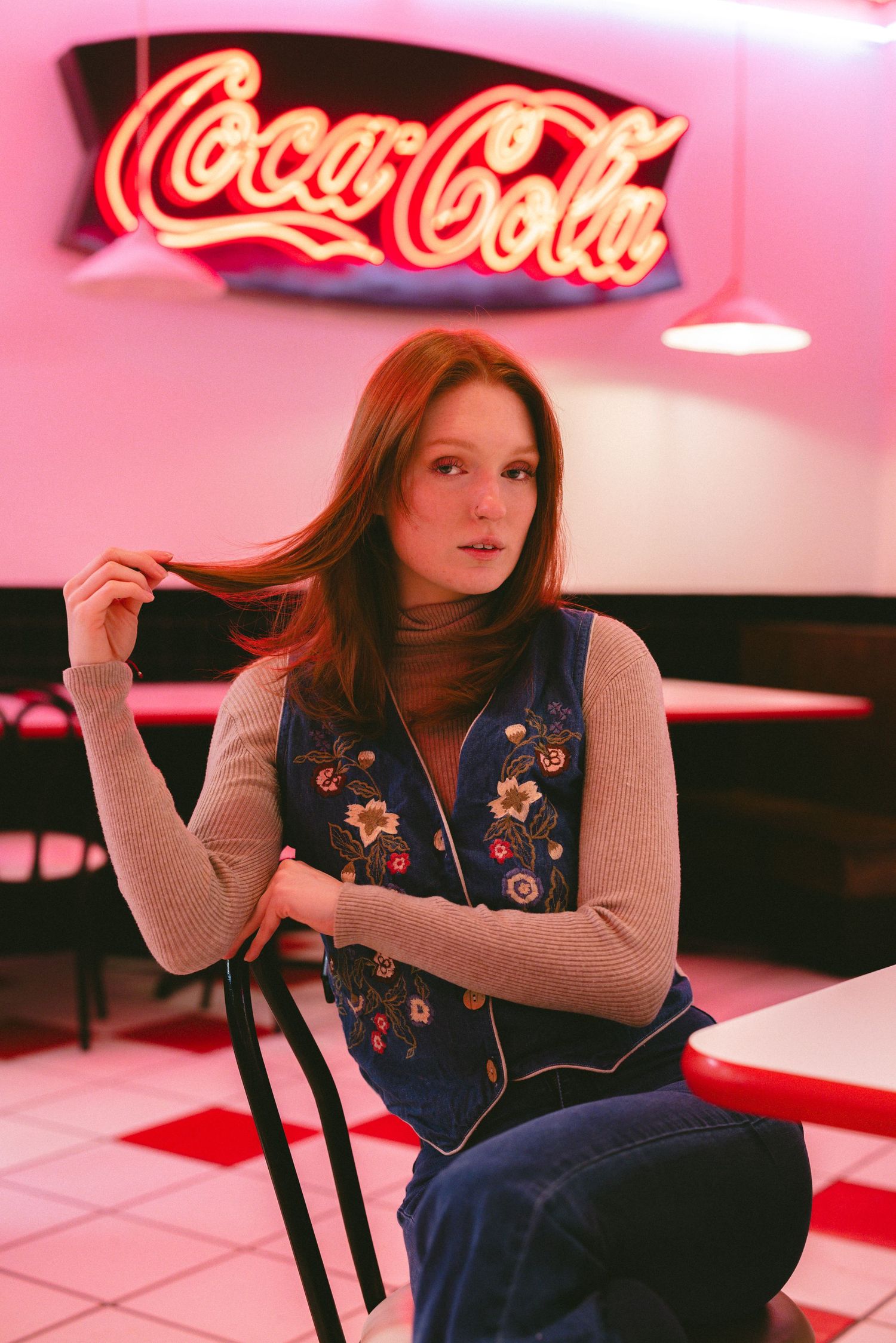 Woman in floral vest lounges in retro diner booth with pink Coca-Cola sign glowing overhead.