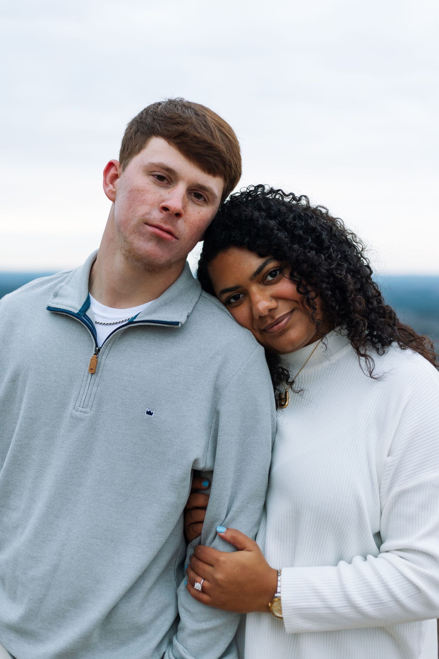 Couple in matching light colored sweaters sharing tender moment against cloudy sky.