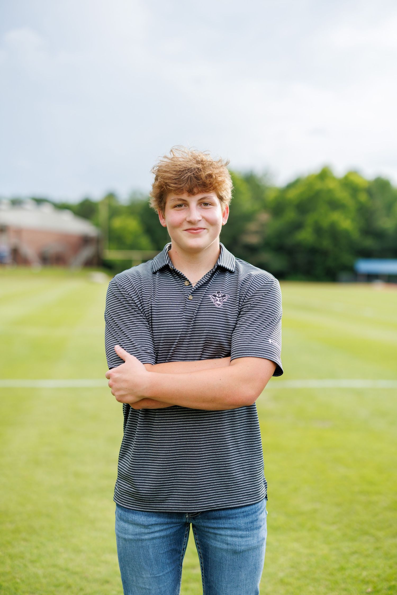 A person in a striped polo shirt and jeans stands with crossed arms in a grassy field on a sunny day.