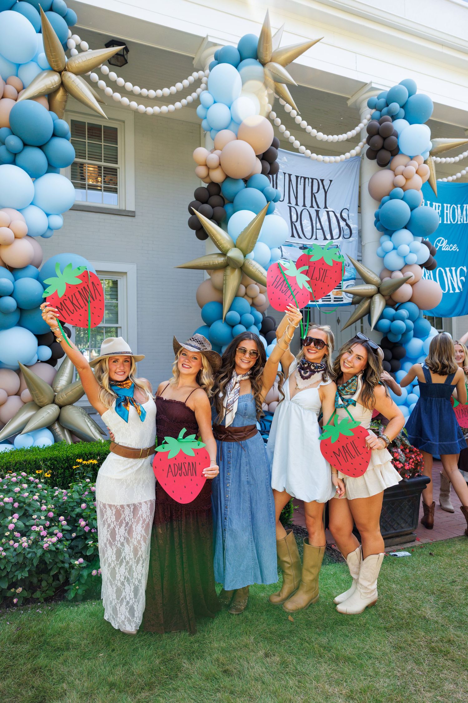 Five women in country western outfits pose with strawberry signs at a Country Roads themed party with balloon arch.