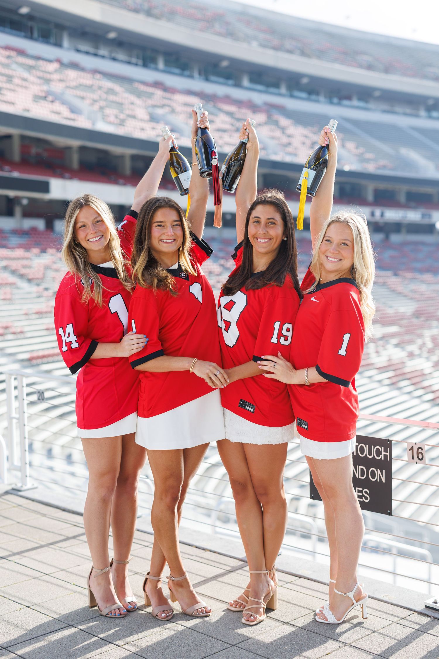 Four women in red sports jerseys pose together in a stadium, raising accessories overhead and smiling.
