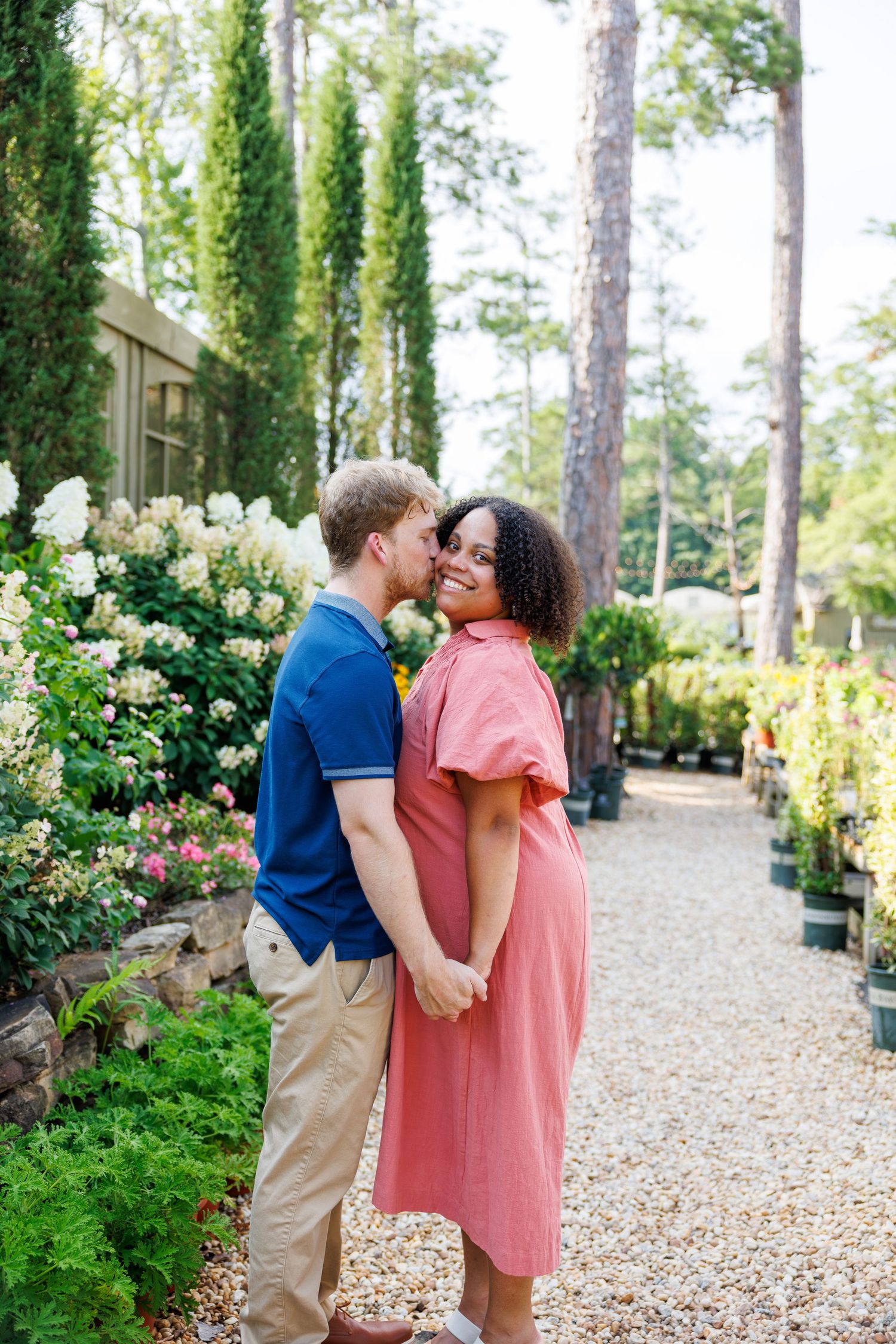 Interracial couple holding hands and sharing a kiss in a lush garden pathway surrounded by blooming flowers.