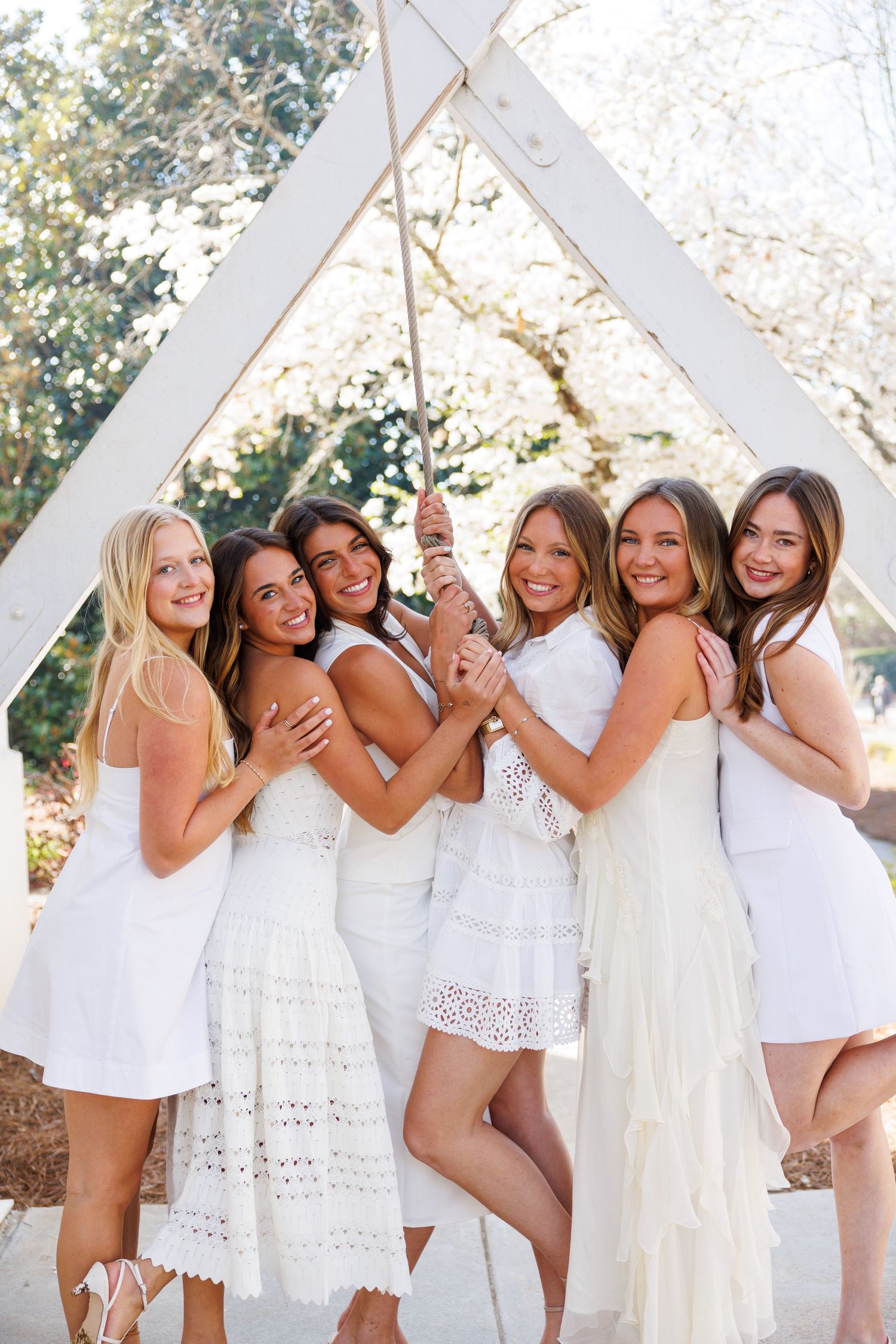 Six women in white dresses pose together under a wooden arch with blooming trees in the background.