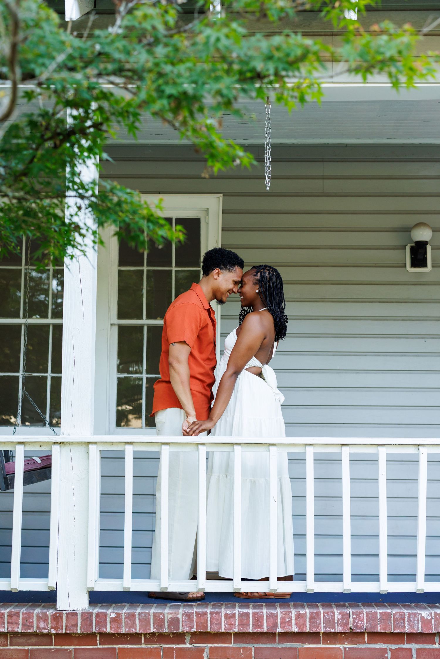 A couple shares a romantic moment on a white porch, the woman in a white dress and man in an orange shirt holding hands.