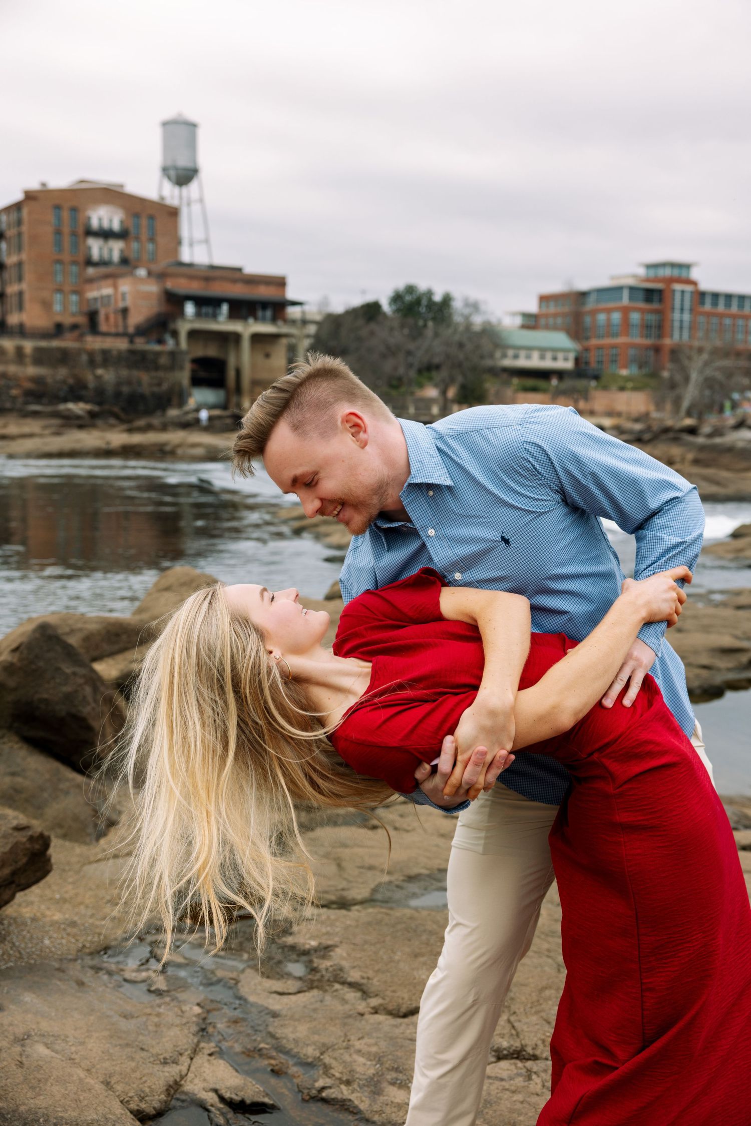 Romantic couple photoshoot on rocky riverbank with city backdrop.
