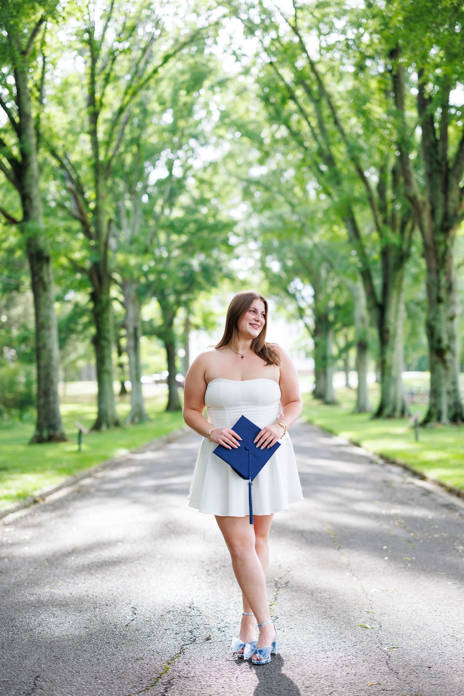 Young woman in white strapless dress holding blue graduation cap on tree-lined path.