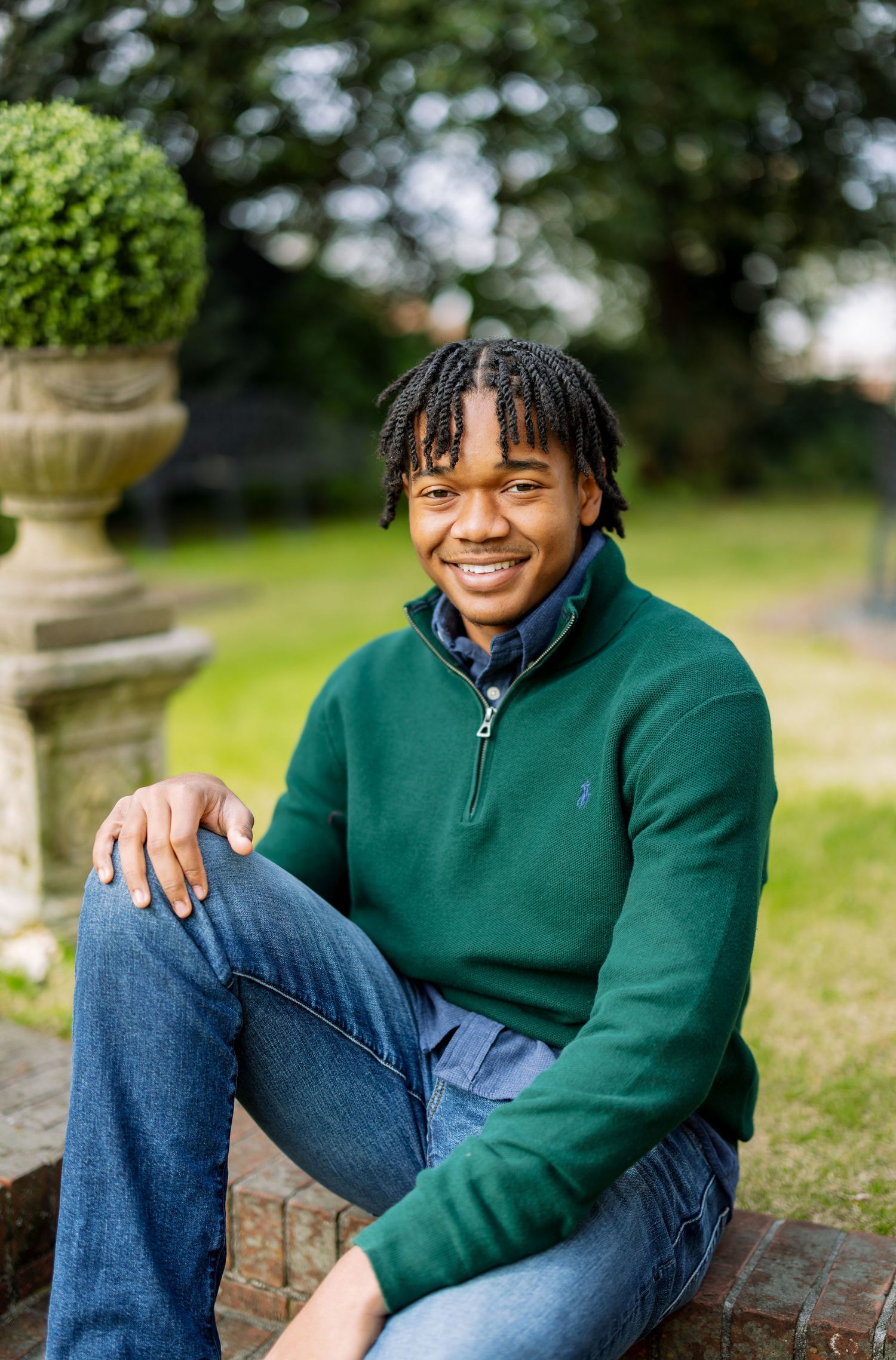 High School Senior in green fleece and jeans poses casually on stone wall outdoors.