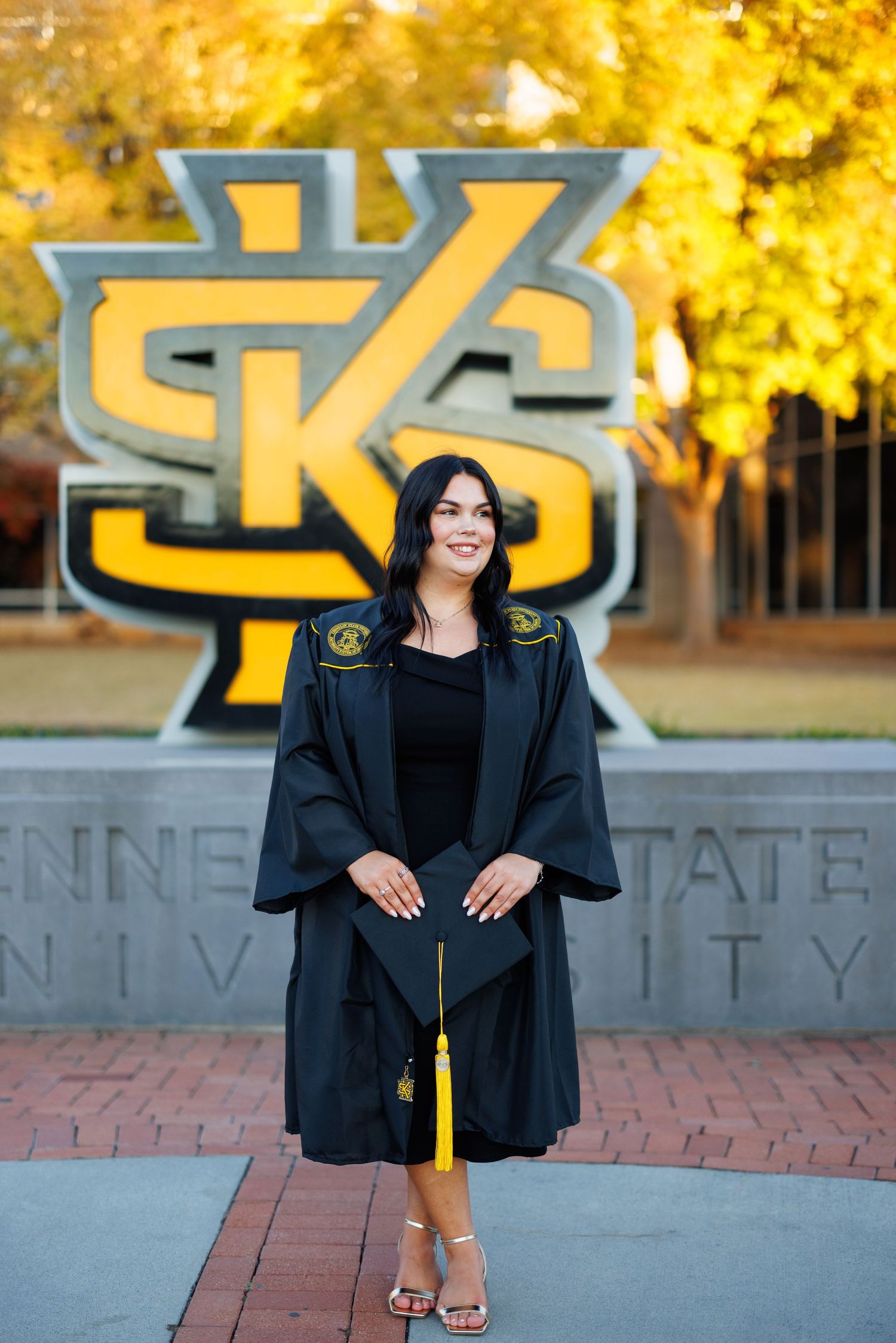 Graduate in traditional black cap and gown poses by university signage.