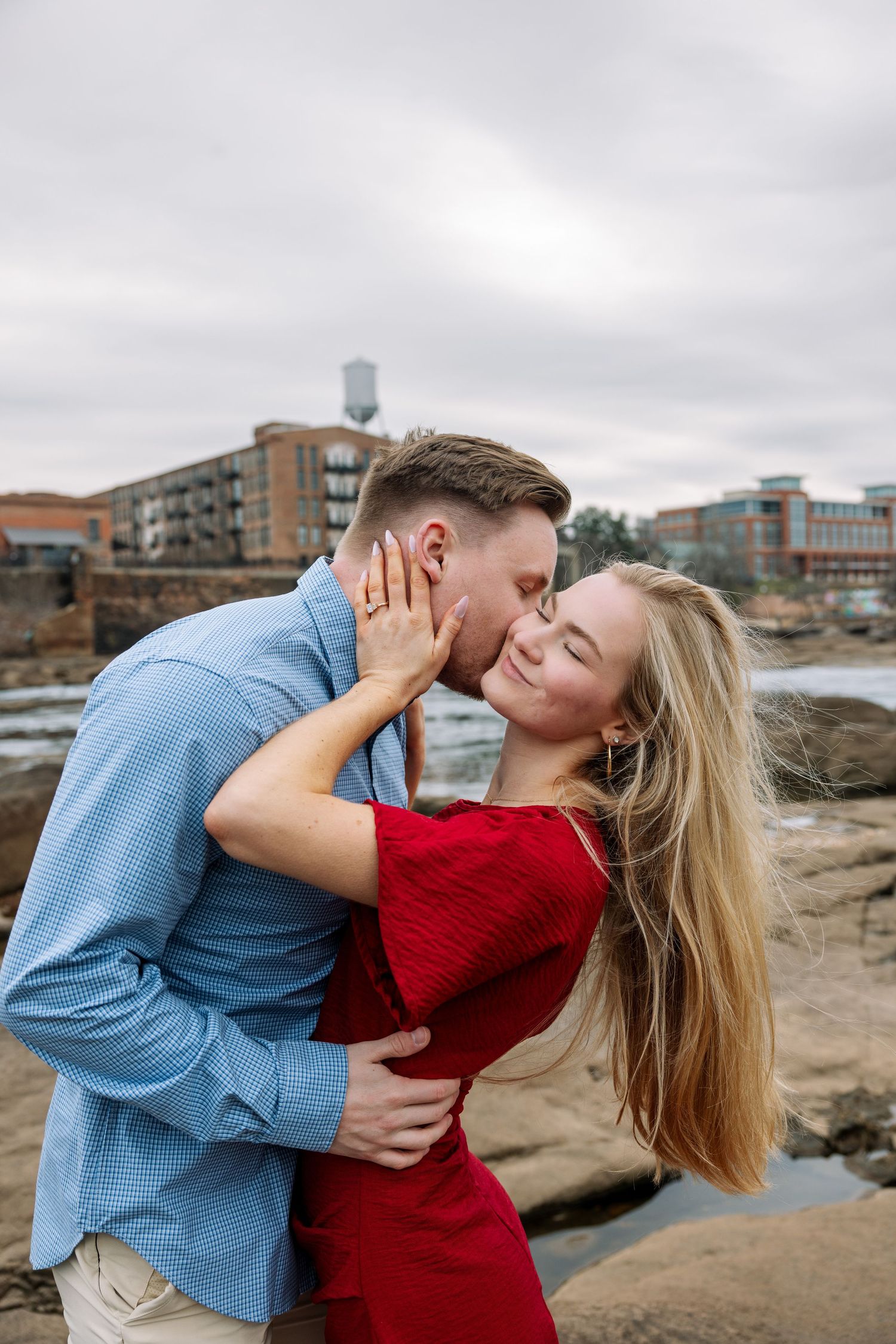 A romantic moment captured between two people on coastal rocks with city buildings in the background.