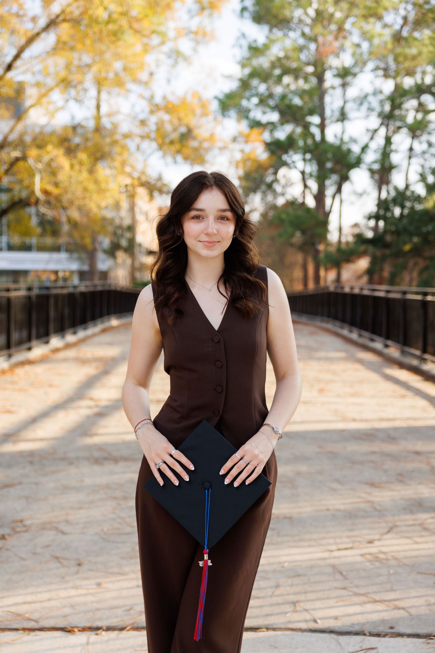 A person in a brown outfit poses outdoors on a wooden walkway surrounded by autumn trees with golden foliage.