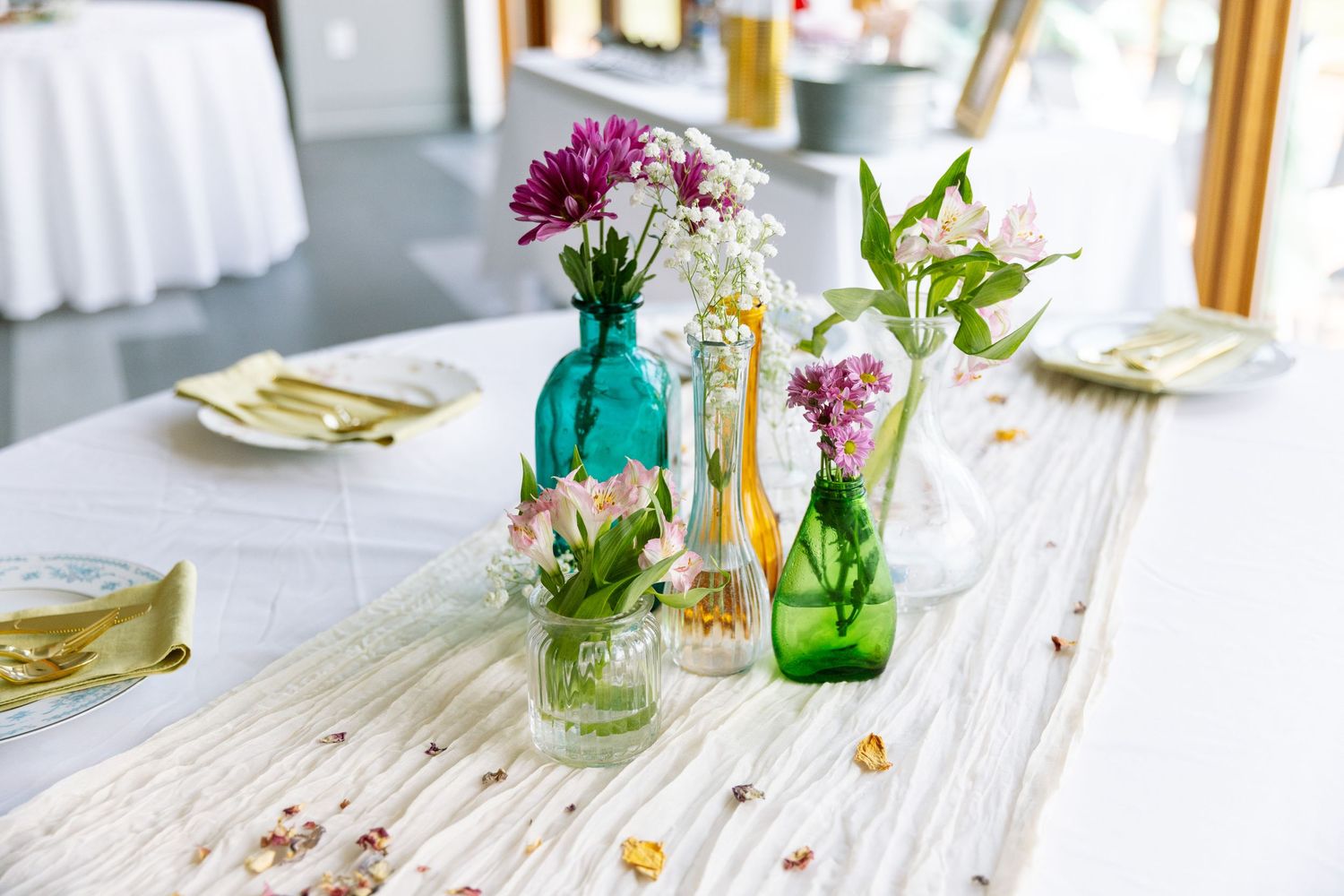 Wedding reception table settings with colorful glass vases and floral centerpieces on white tablecloths.