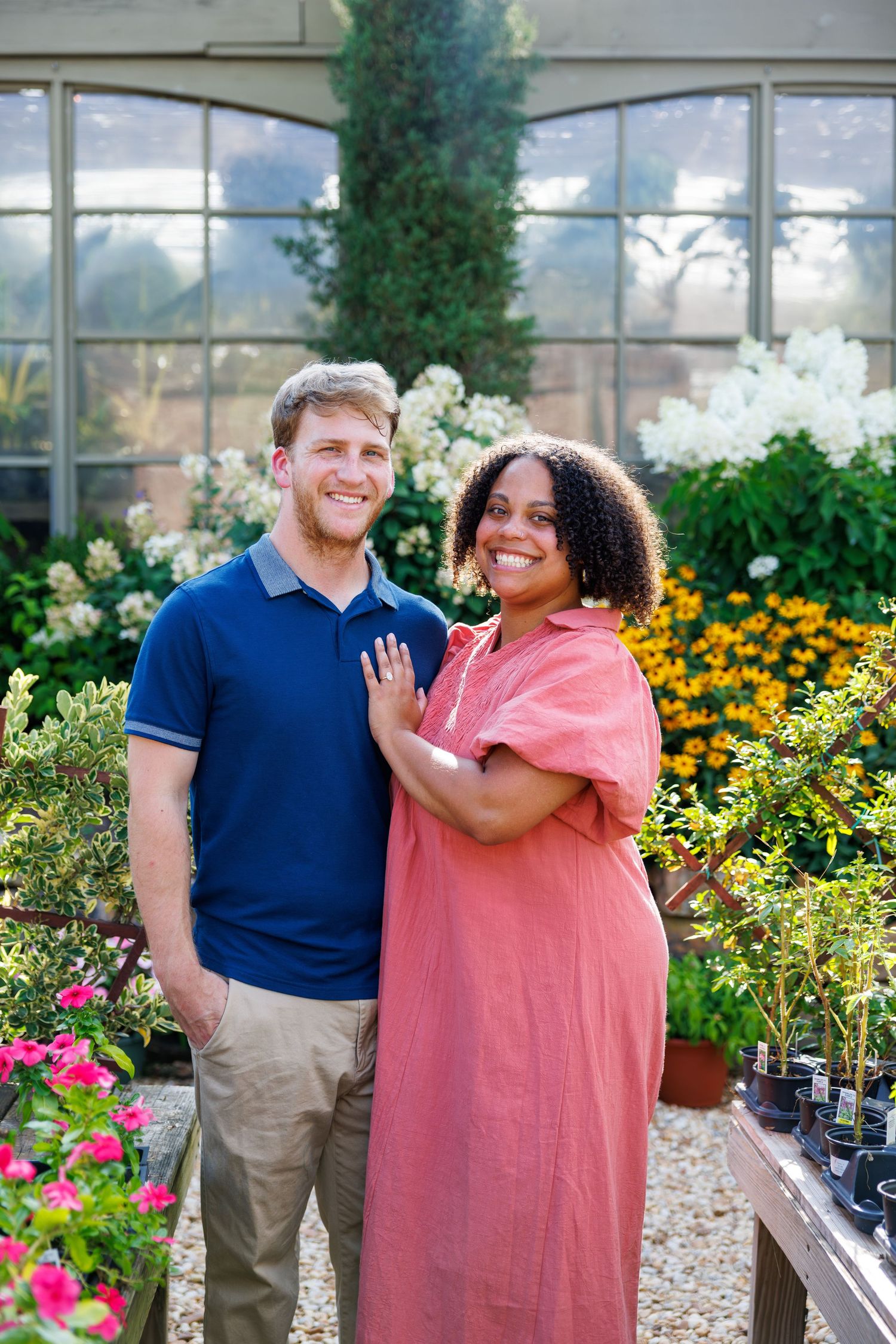 A smiling interracial couple poses together among colorful flowers in a lush greenhouse garden setting.