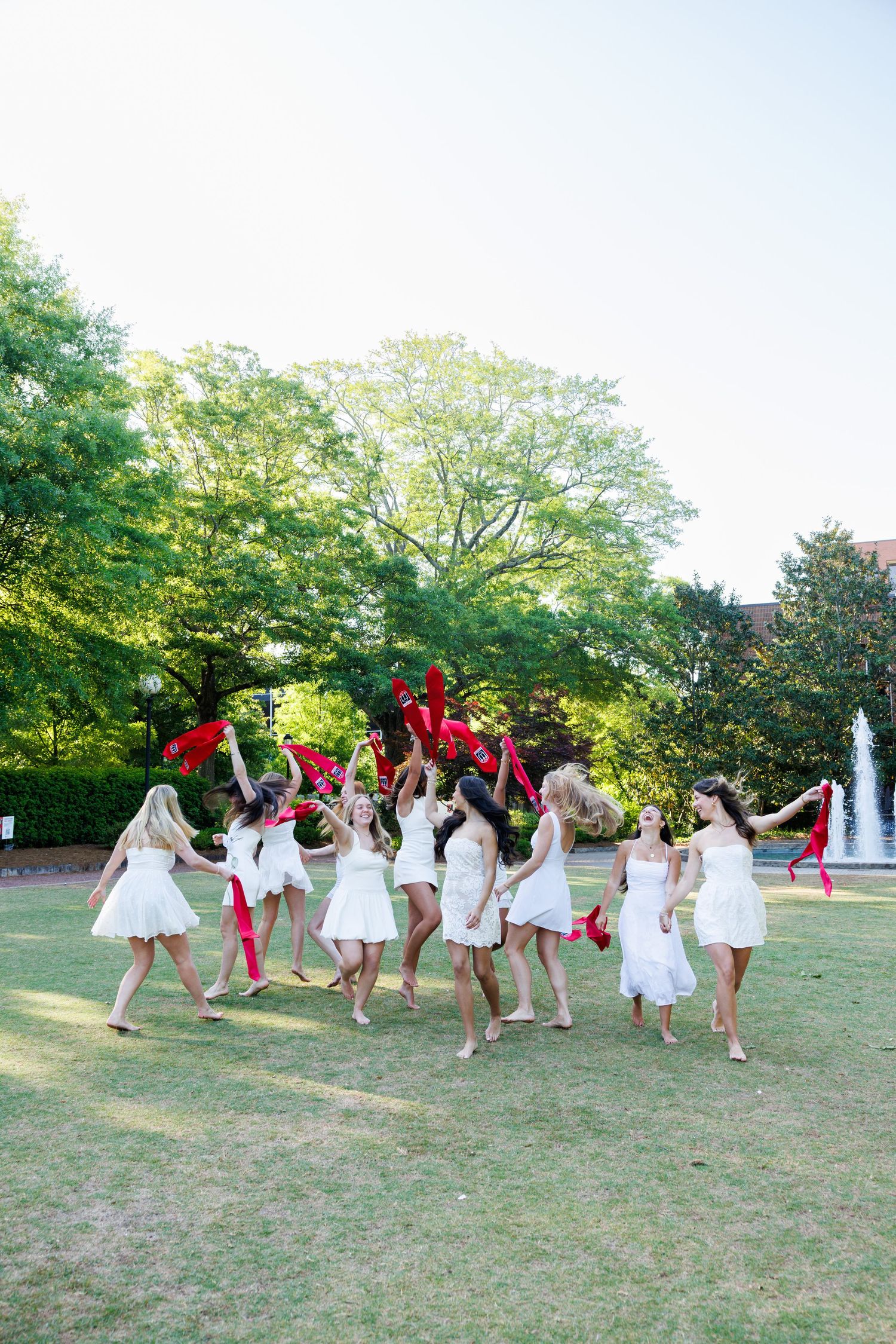 Group of women in white dresses dancing outdoors with red ribbons on a sunny day near a fountain.