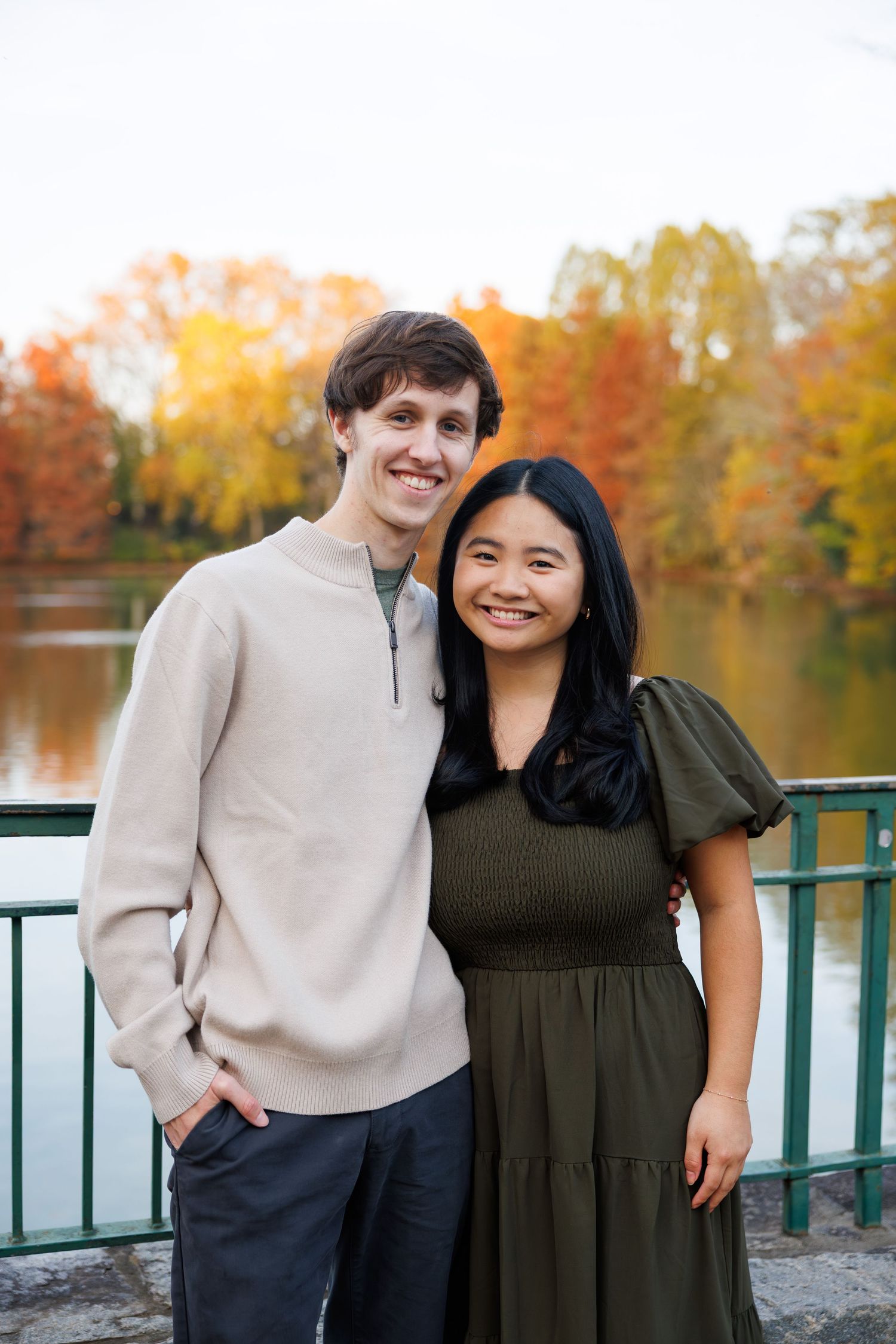 Couple posing together by a lake during autumn with colorful fall foliage in background.