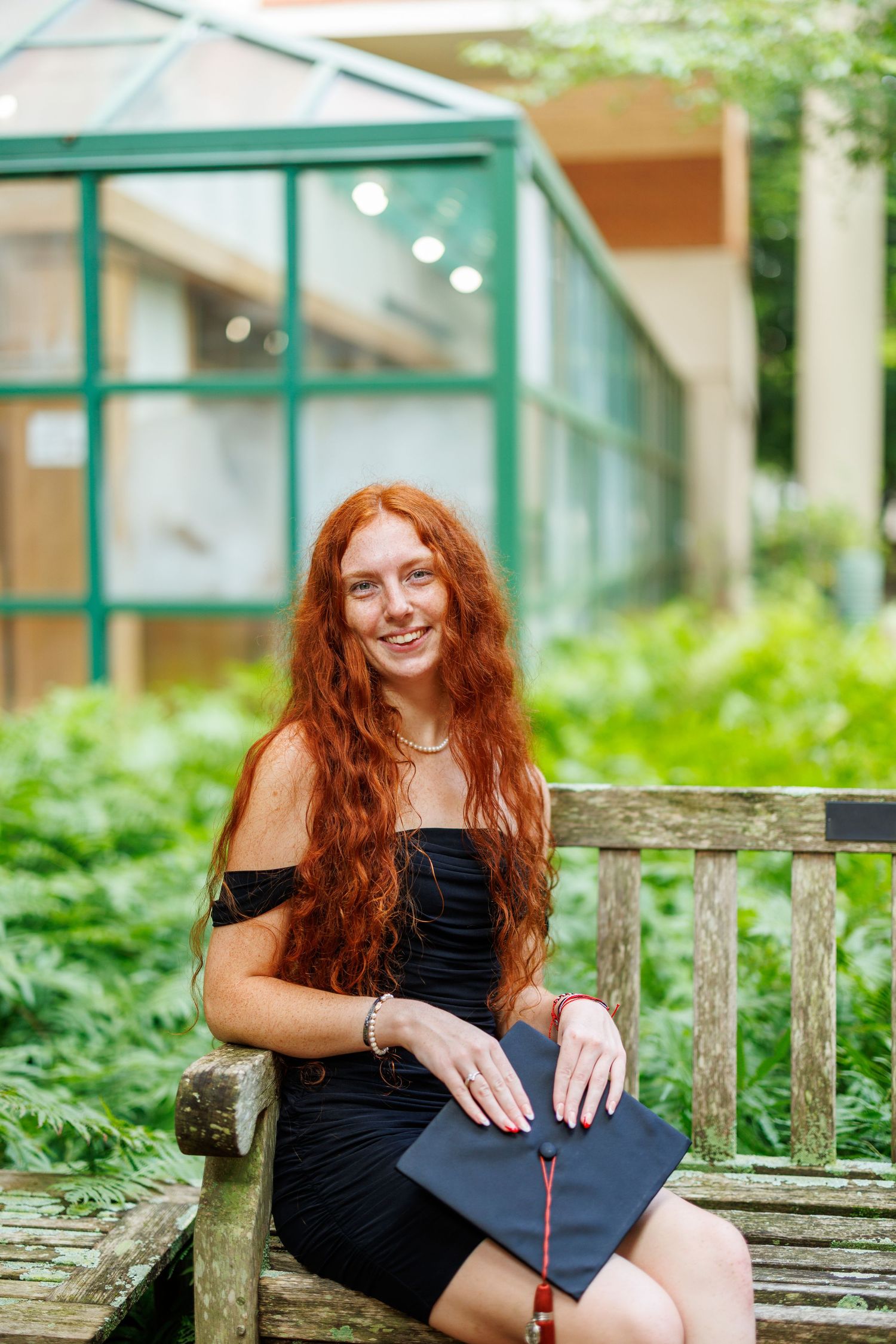 A person with long red hair poses outdoors in a black dress on a rustic wooden bench near lush greenery.