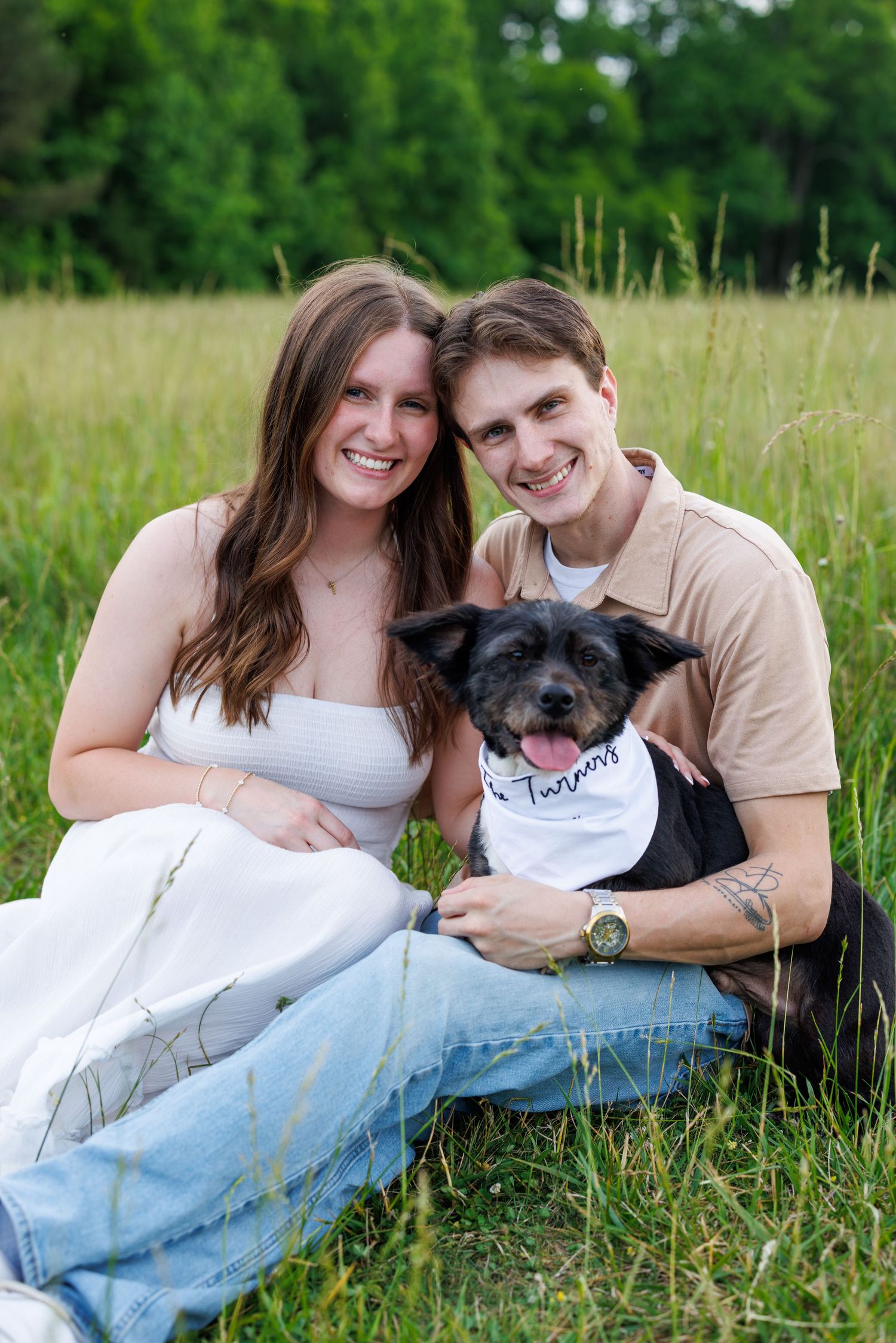 A smiling couple sits in a grassy field with their black dog wearing a bandana, enjoying a sunny outdoor portrait.
