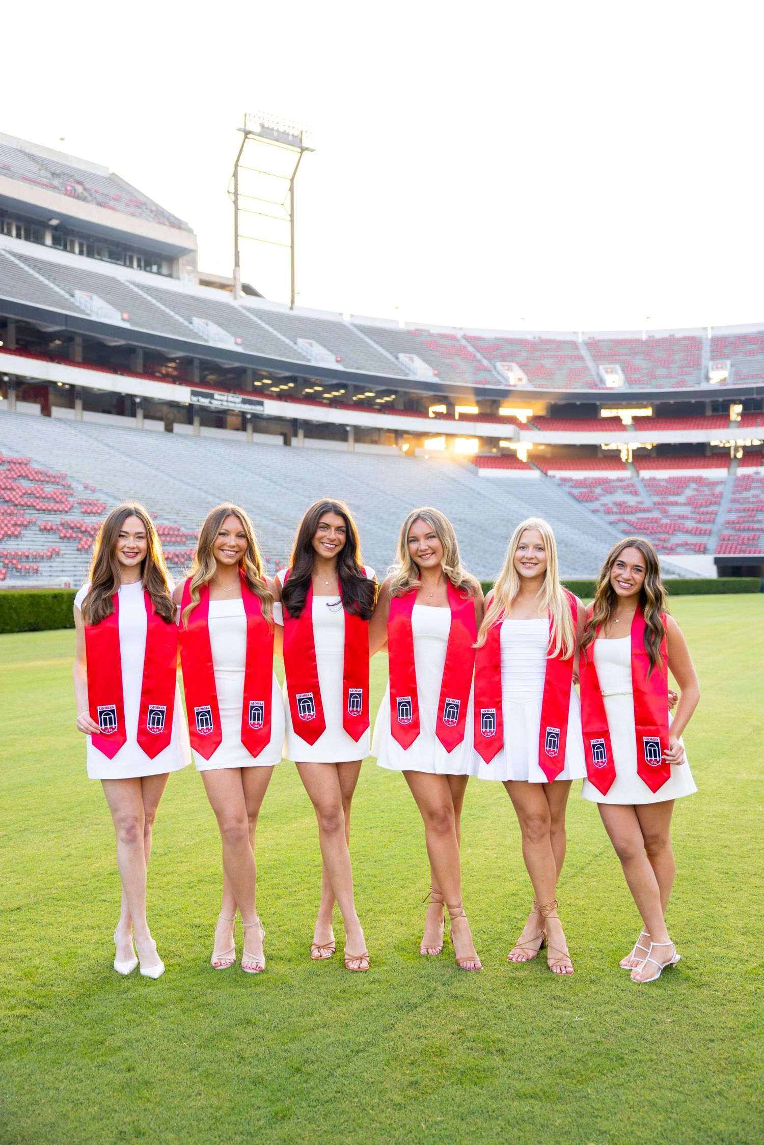 Six young women in white dresses and red graduation stoles pose on a football field stadium.