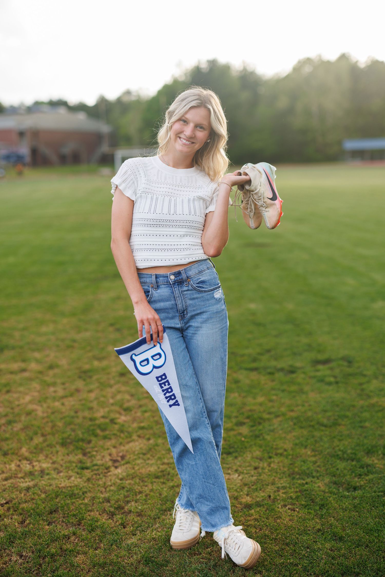 Smiling blonde woman holding Berry College pennant and shoes, wearing white crop top and jeans on green field.