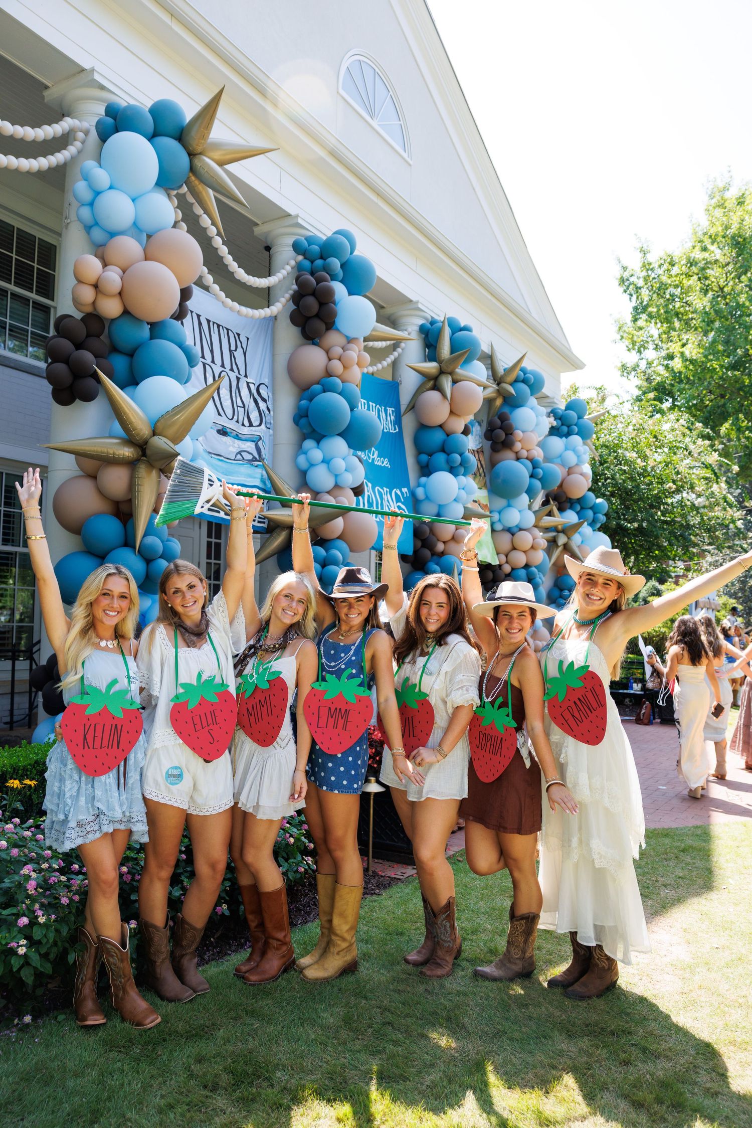 Group of women in cowboy boots posing with strawberry signs at a festive balloon-decorated outdoor event.