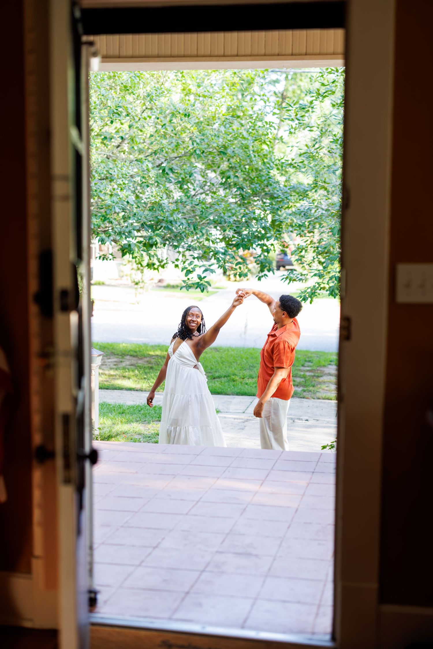 Couple dancing outdoors, woman in white dress and man in orange shirt, viewed through an open doorway.