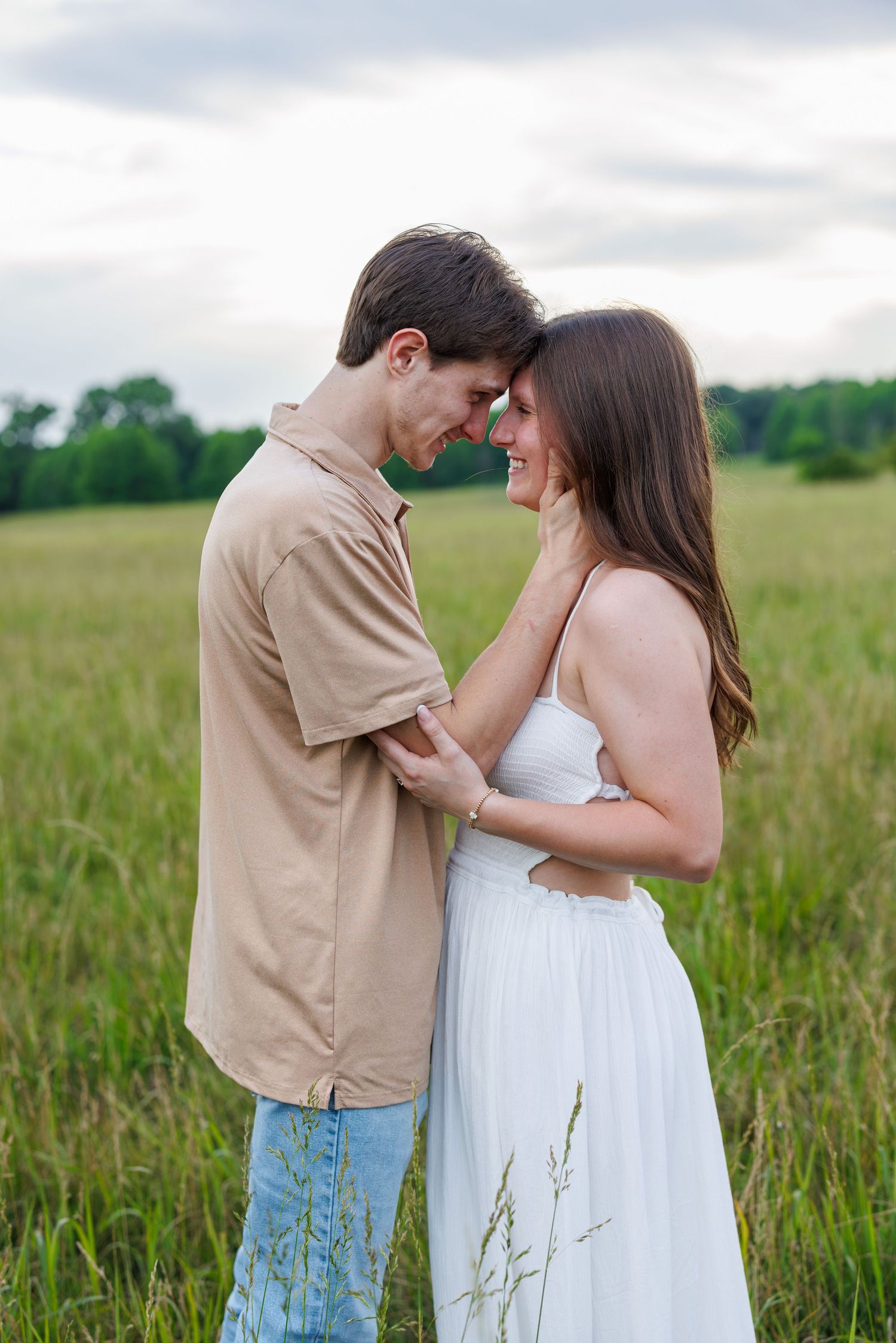 Romantic couple touching foreheads in a green field, woman in white dress and man in beige shirt sharing a tender moment.