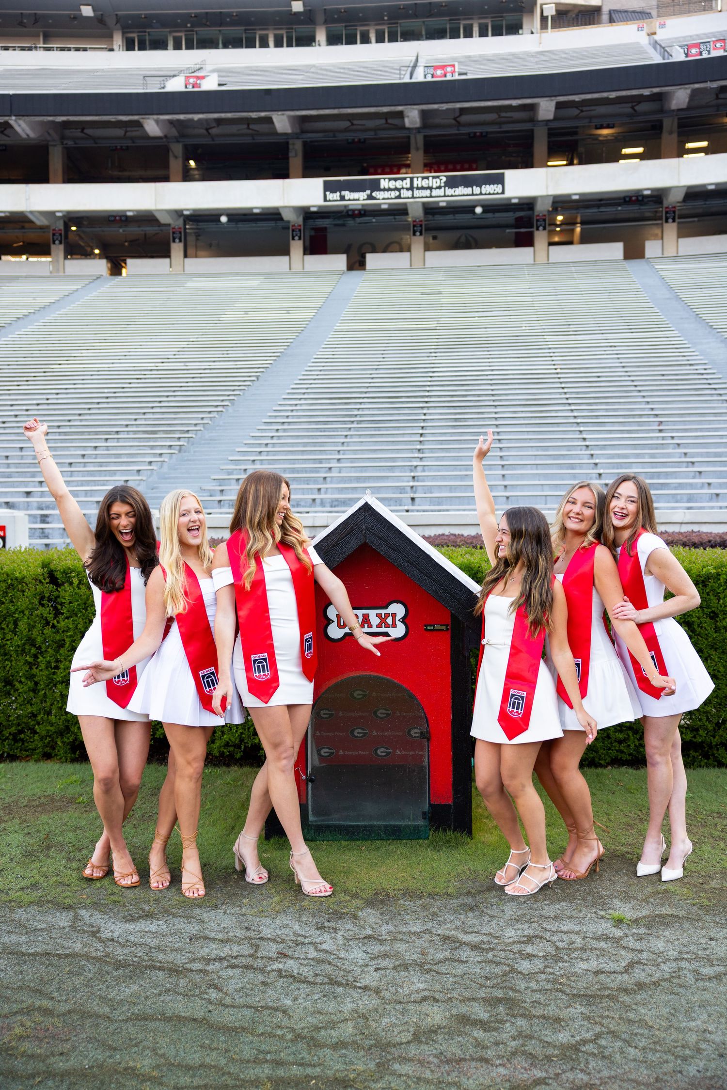 Six women in white dresses and red graduation sashes pose cheerfully in a stadium near a small decorative doghouse.