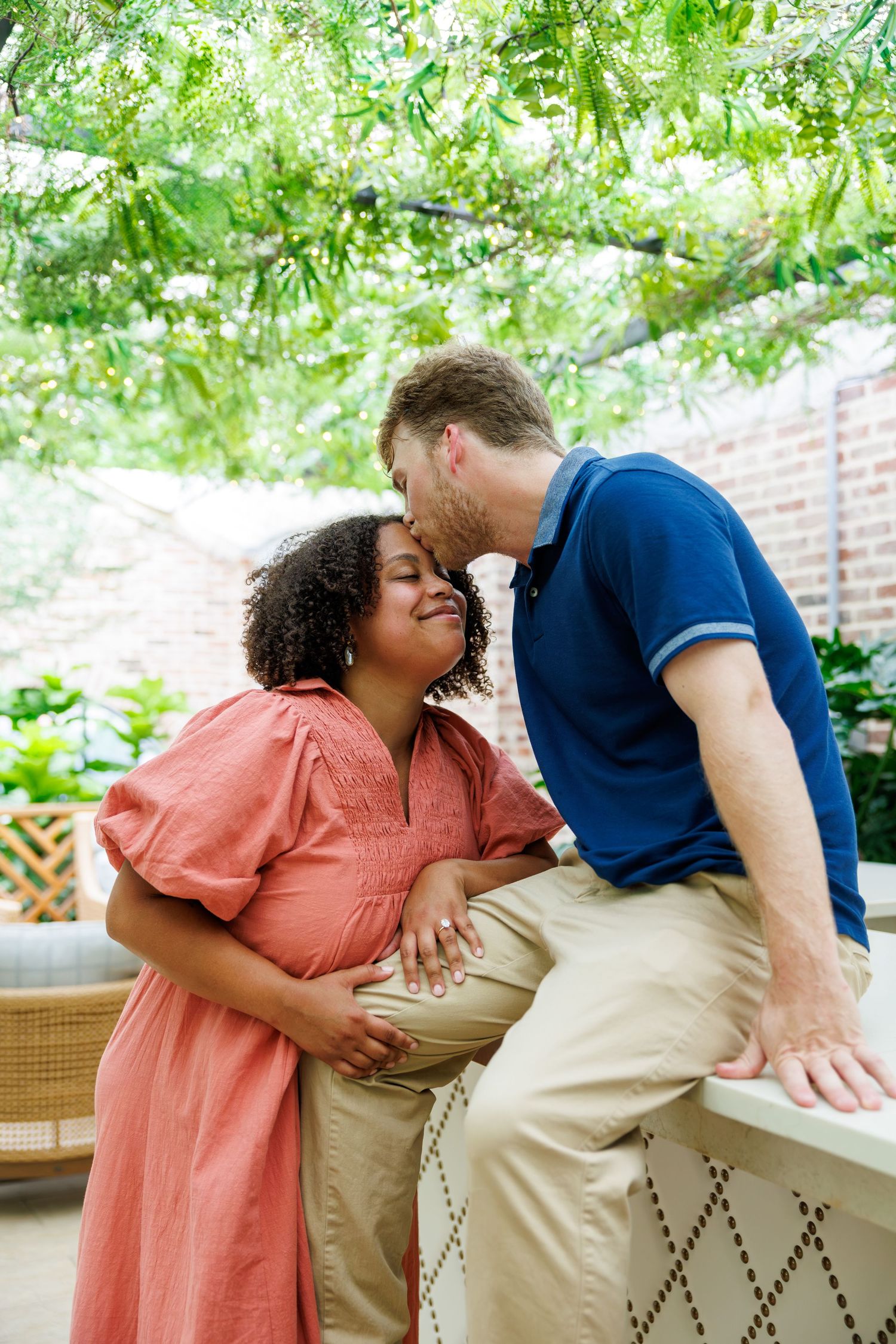 Pregnant interracial couple sharing a tender kiss under green vine canopy, man gently touching baby bump.