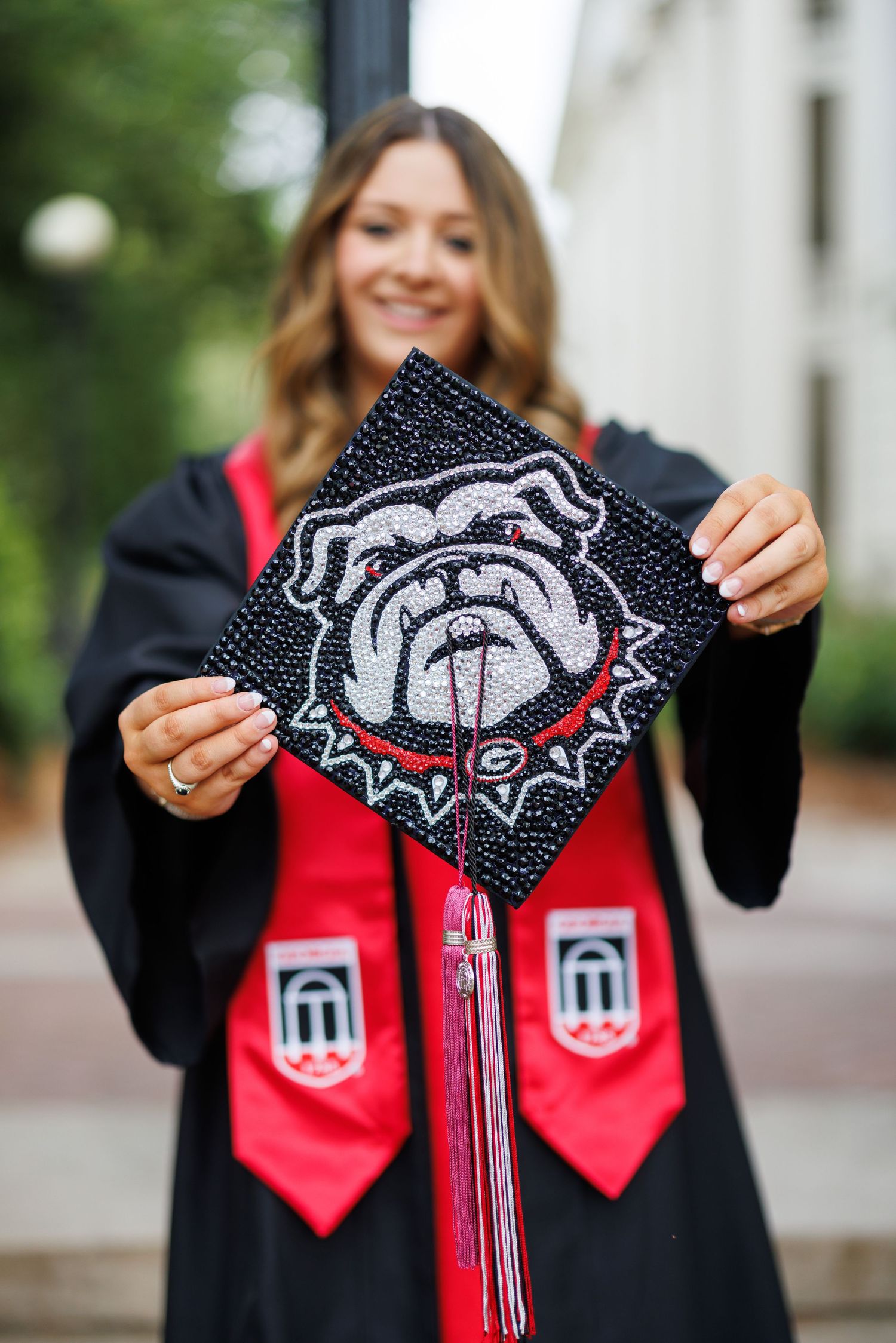 Graduate in black cap and gown holds decorated graduation cap with University of Georgia bulldog mascot design.