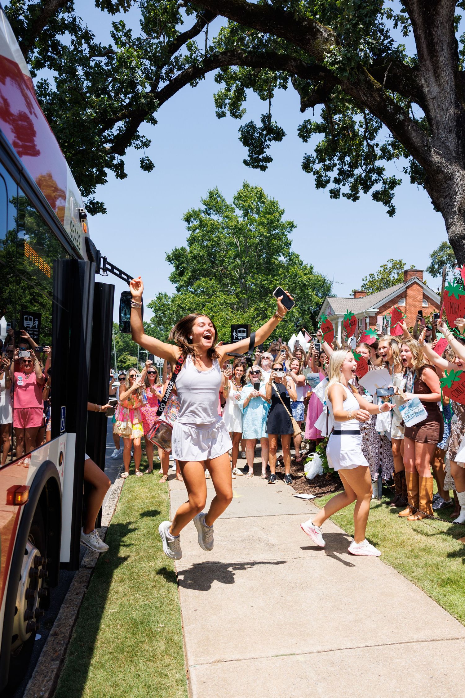 Two young women in white outfits celebrate joyfully outdoors near a bus, surrounded by cheering crowd on a sunny day.