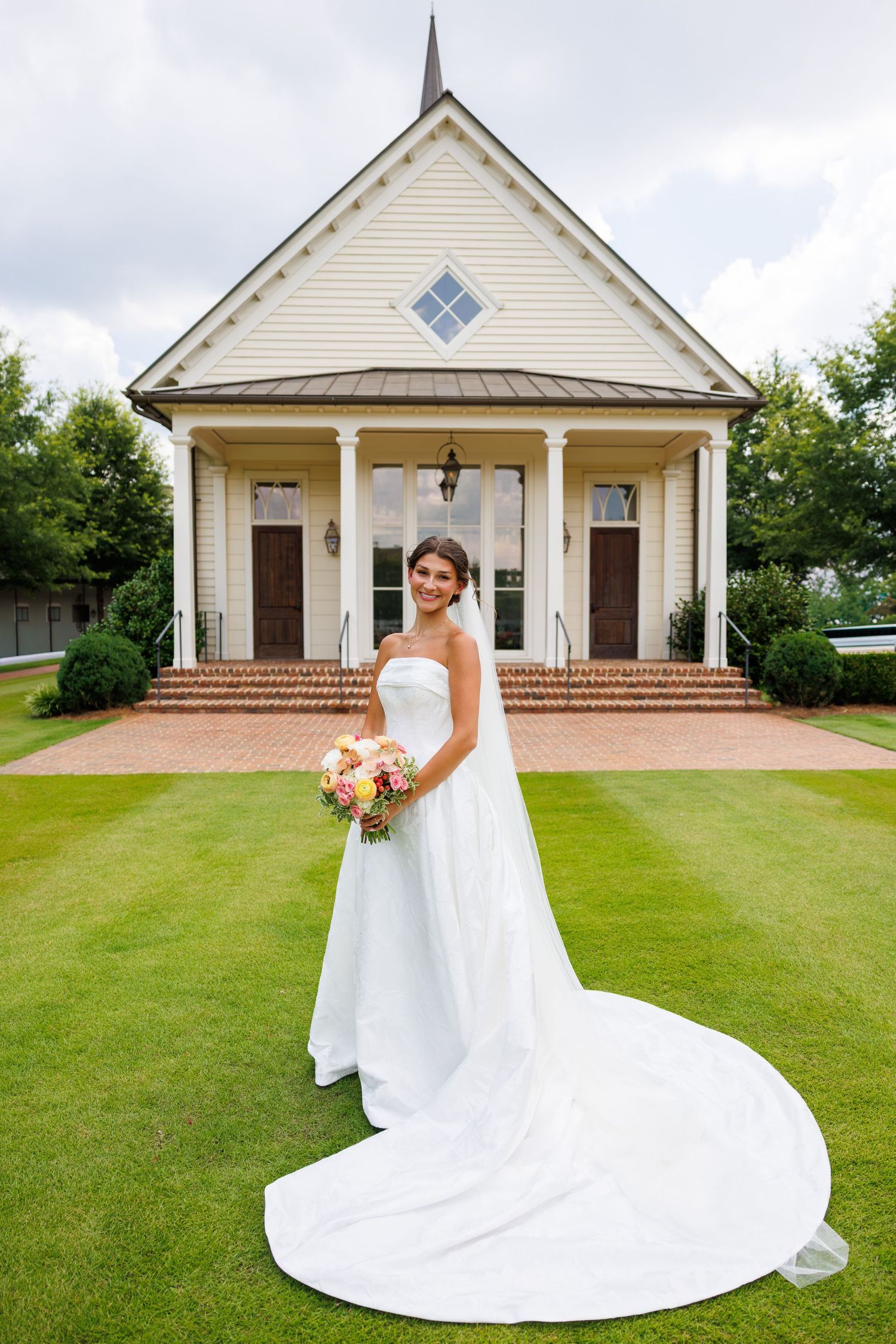 Bride in strapless white gown with long train holds bouquet on green lawn before charming white chapel.