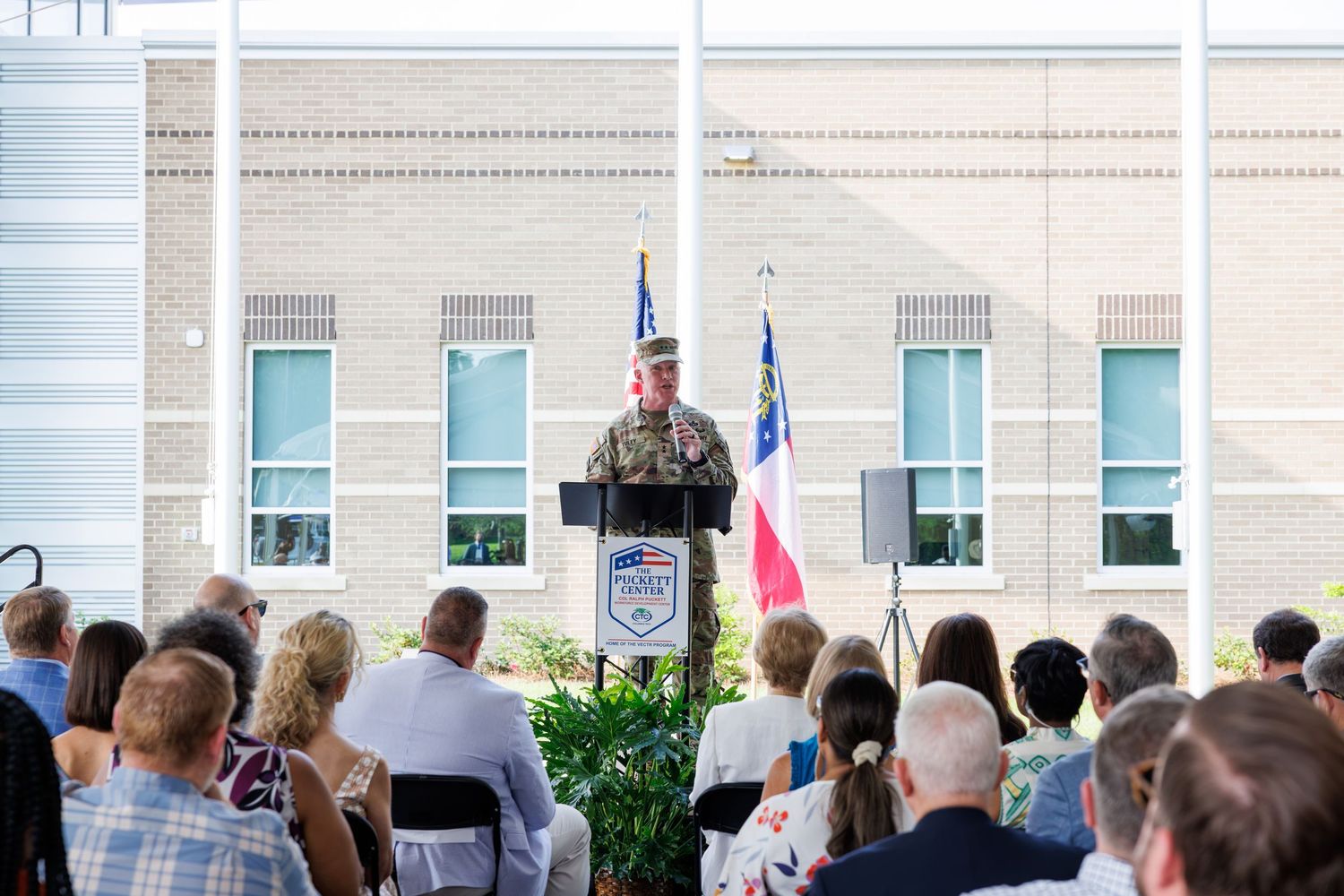 Military officer speaks at outdoor podium during official ceremony with audience and flags in background.