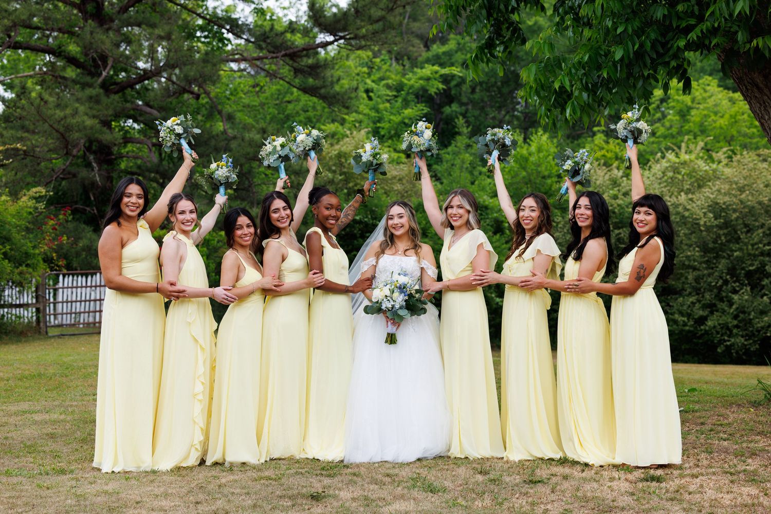 Bride in white gown poses with eight bridesmaids in yellow dresses holding bouquets aloft outdoors.