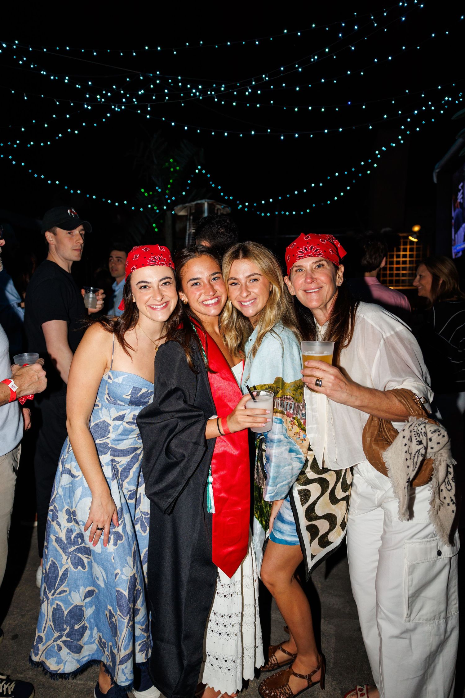 Four women celebrating a graduation outdoors at night, with string lights overhead, one wearing cap and gown.