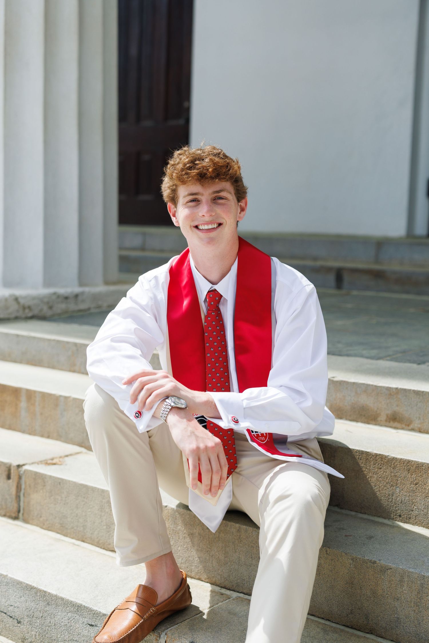Smiling young man in white shirt, red tie, and graduation stole sitting on stone steps outdoors.