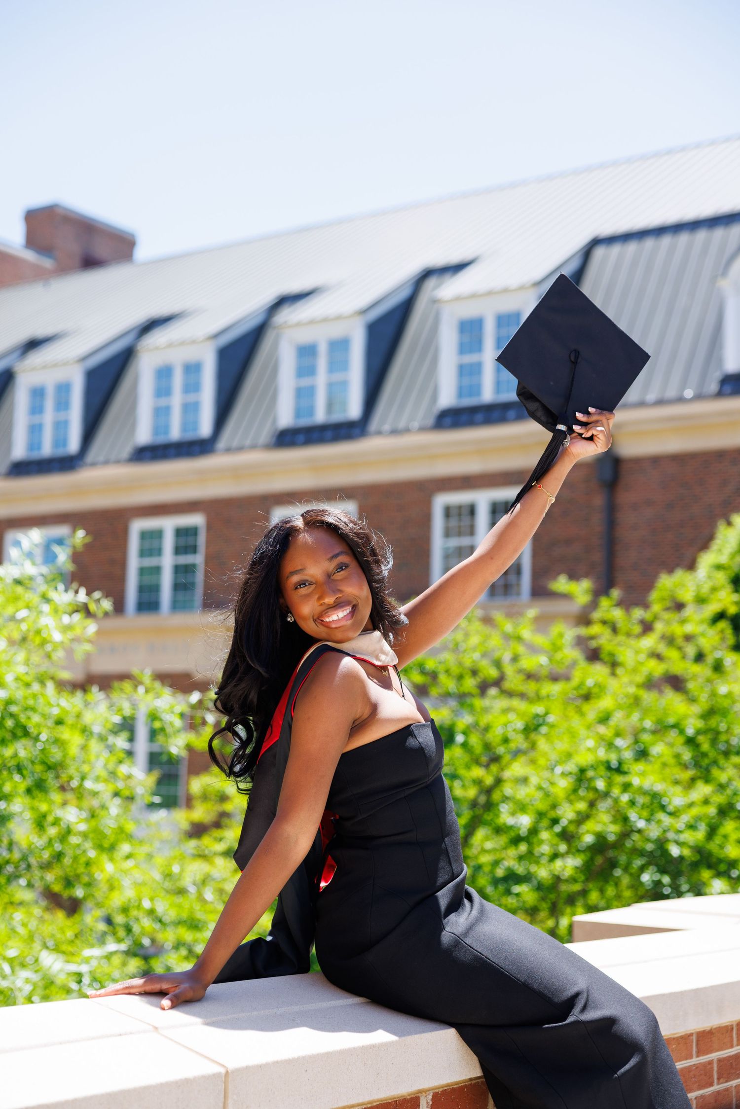 Smiling Black female graduate in cap and gown sitting outdoors, raising her mortarboard in celebration on a sunny day.
