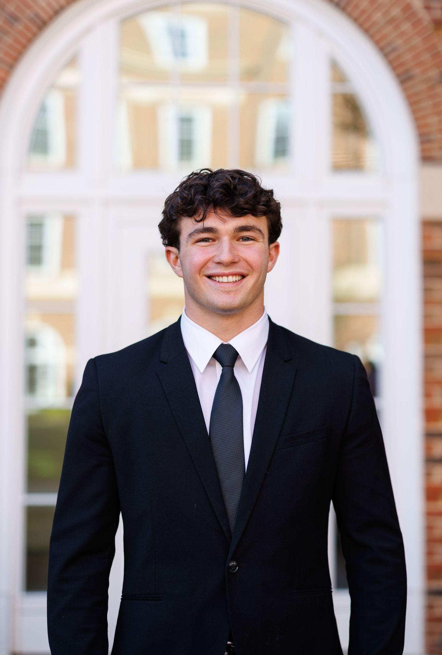 Young man in black suit and tie smiling in front of arched building entrance.