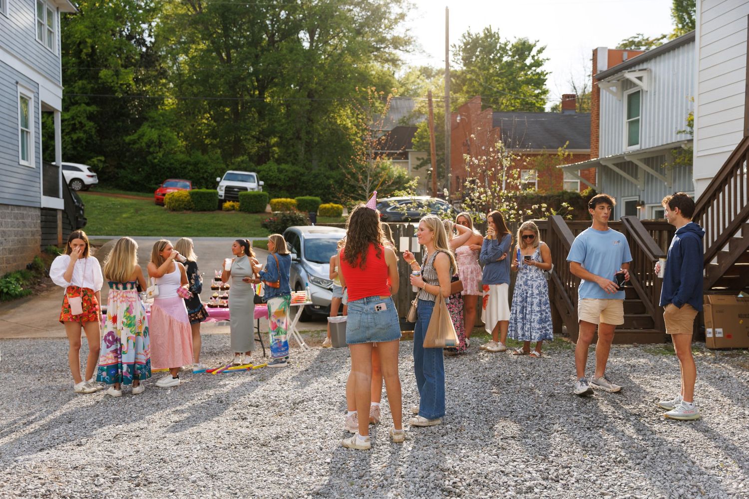 Group of teens and children gathered on a gravel driveway between houses during a sunny summer evening outdoor party.
