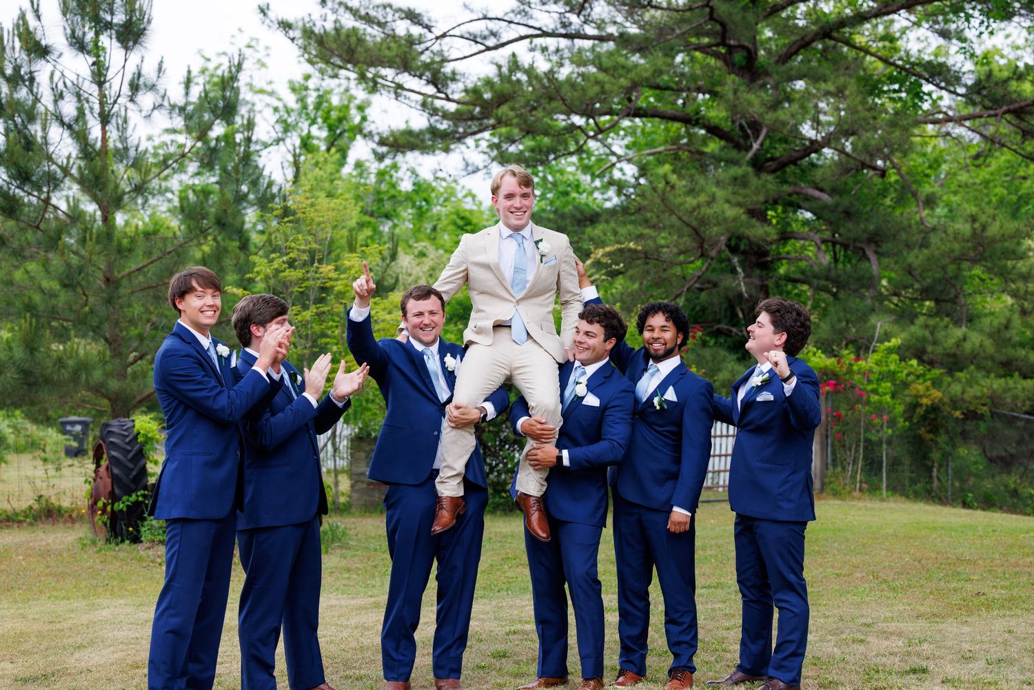Groomsmen in navy suits joyfully tossing the groom in the air outdoors before the wedding ceremony.