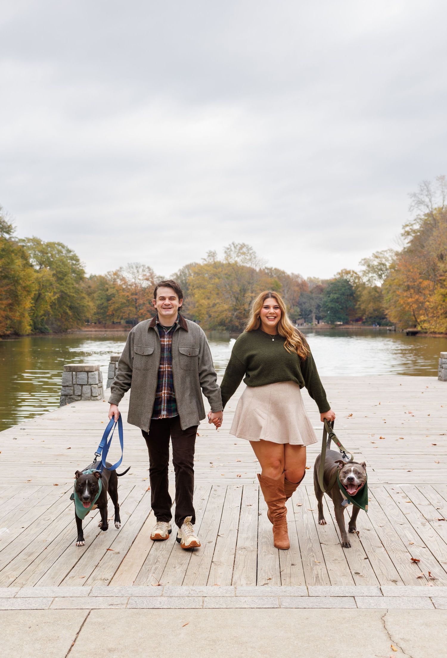A couple walks their two dogs along a wooden dock by an autumn lake.