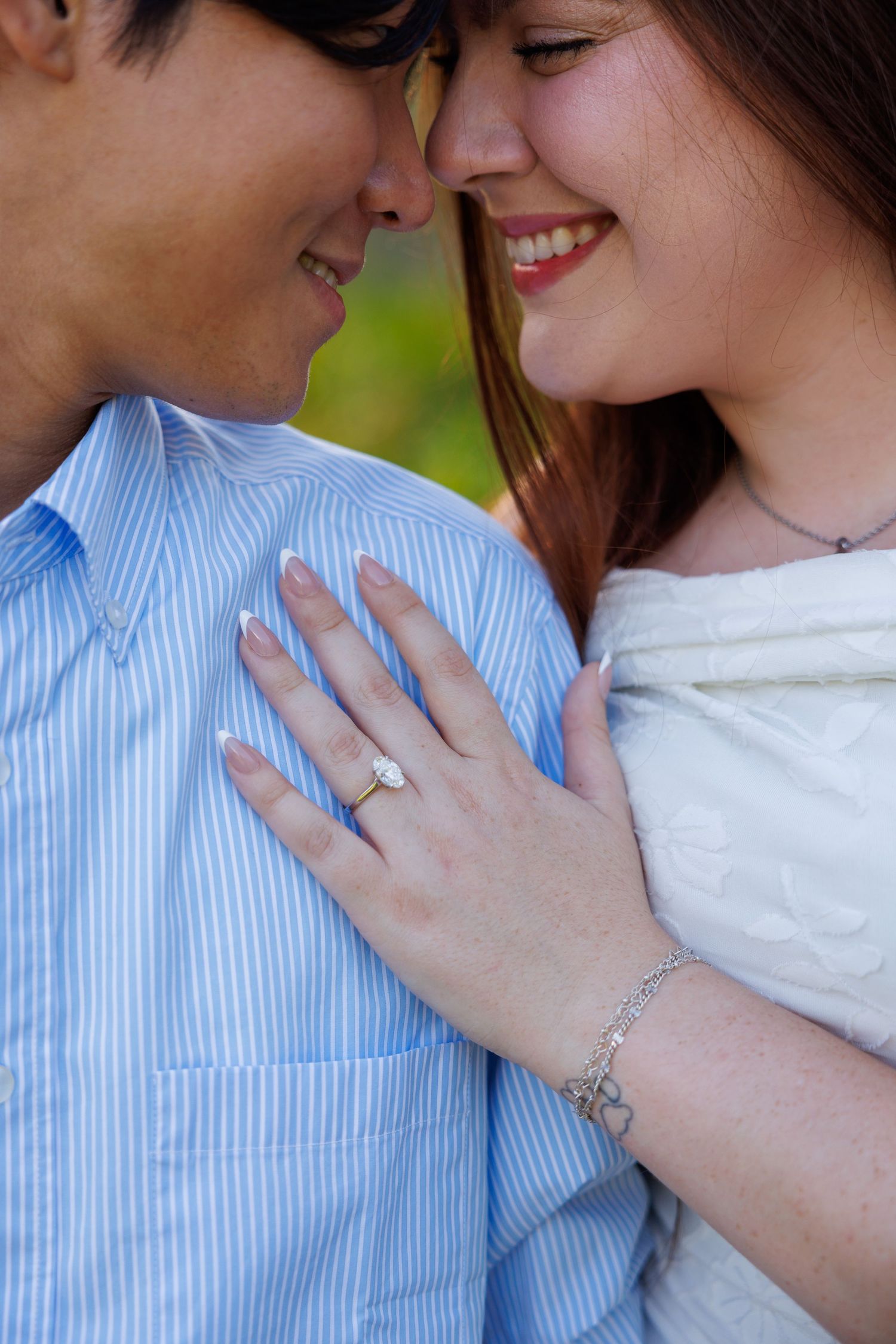 Happy couple smiling face-to-face, woman wearing diamond engagement ring and bracelet, hand resting on man's blue striped shirt.