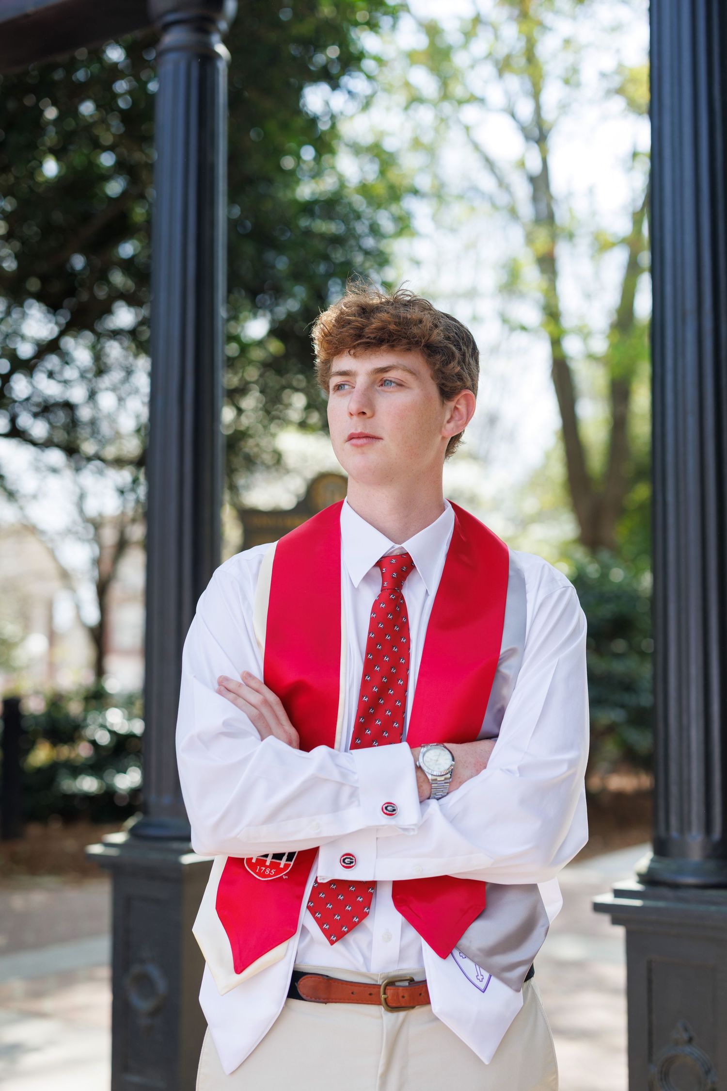 Young man in white dress shirt, red tie, and graduation stole stands with arms crossed outdoors.