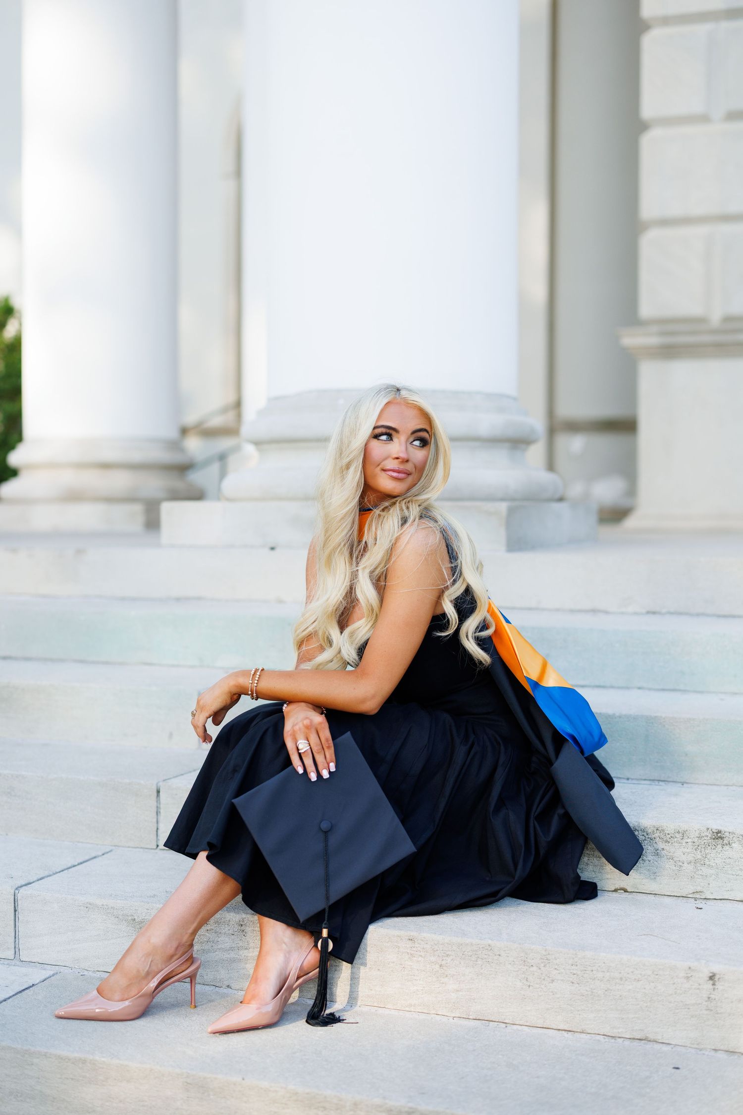 Blonde woman in black graduation gown and heels sits on stone steps holding cap with colorful hood.