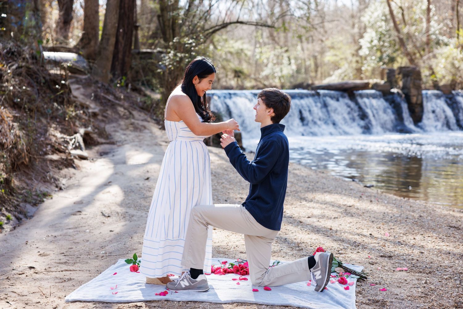 A romantic picnic scene by a flowing river with a couple enjoying wine and snacks on a sunny afternoon.