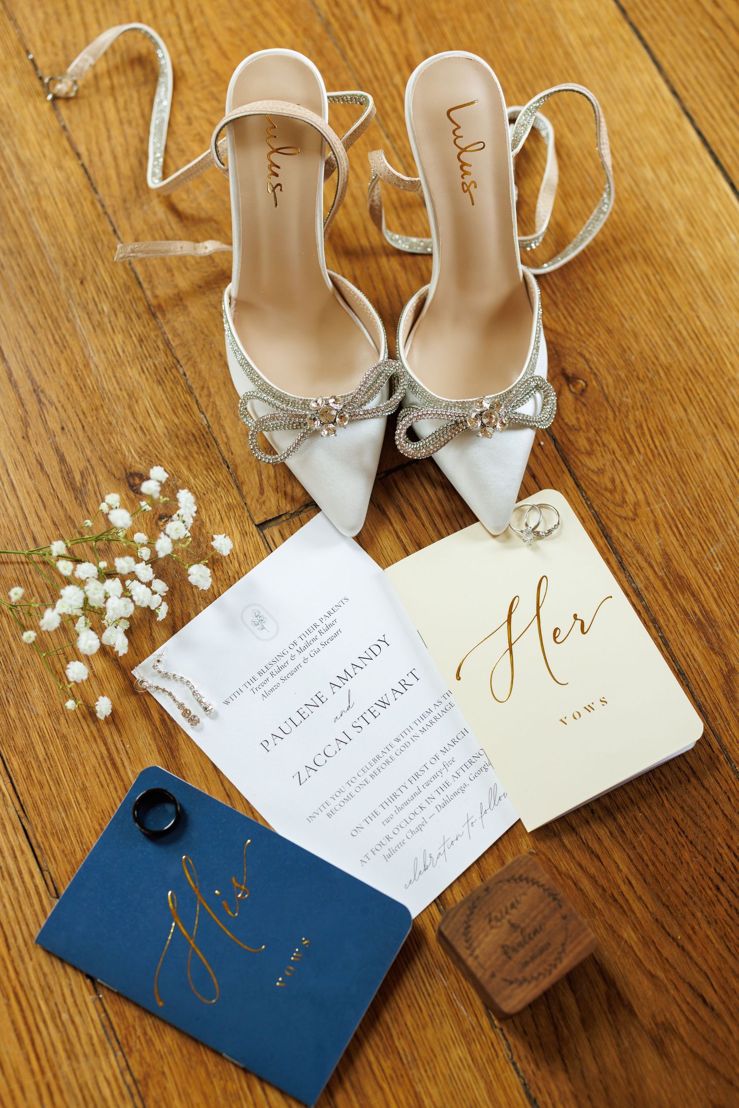Elegant bridal flat lay featuring white bow heels, wedding invitations, His and Her vow books, and baby's breath on wood floor.